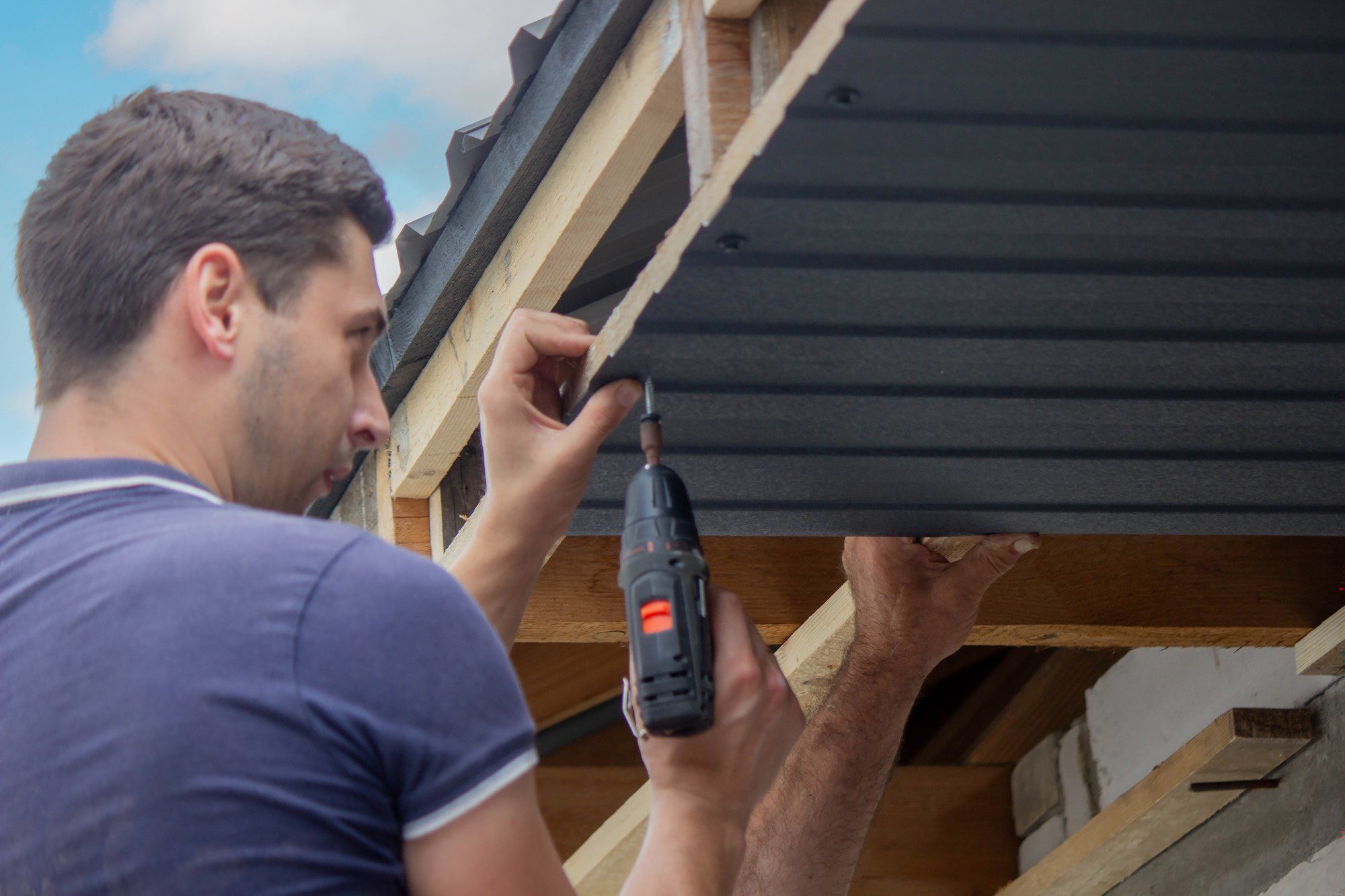 The image shows a man engaging in some kind of construction or home improvement work. He is using a cordless drill to fasten material, which appears to be a piece of roofing or siding, to what seems to be part of a house or a building structure. The man is focused on his task, securing the material with screws or nails, and it appears that someone else is assisting him by holding the material in place. The setting suggests an outdoor environment given the presence of the sky in the background. Overall, the scene depicts a moment of hands-on work, likely related to building or repairing a part of a home or structure.