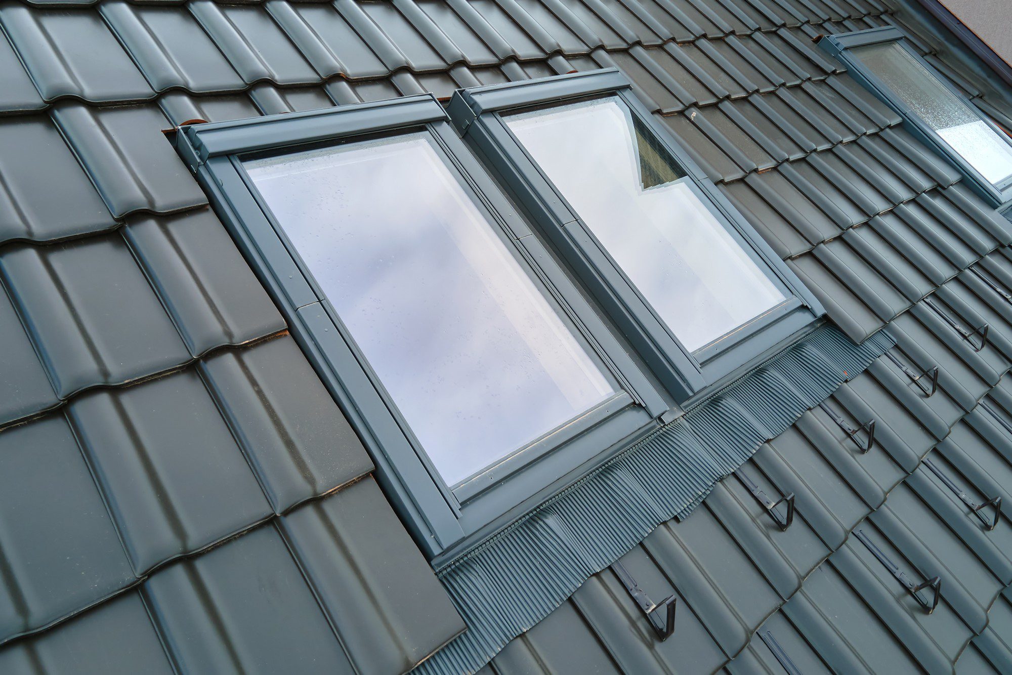 This image shows a close-up view of a roof with two skylight windows. The roof is covered with interlocking metal tiles, which are common for their durability and weather resistance. The skylight windows are framed with a metal or possibly PVC material, which is flush with the roofing material to provide a seamless look and ensure waterproofing. The windows also appear to have some water droplets on them, suggesting recent rain or condensation. The angle of the photo emphasizes the pattern and texture of the roof tiles, as well as the design and installation of the skylight windows. There is a slight reflection visible on the glass, but it's unclear what it is reflecting due to the overcast sky.