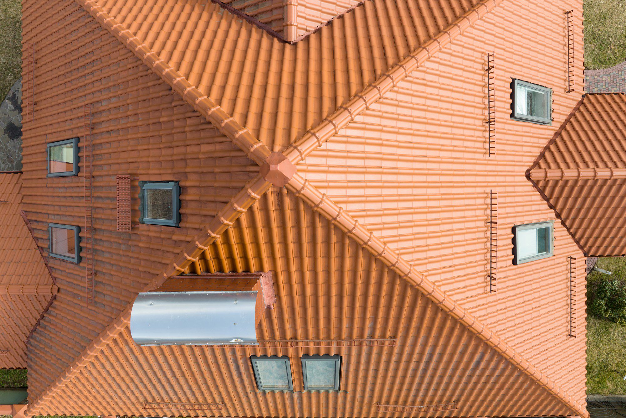 The image displays a top-down aerial view of a series of pitched roofs joined together in a complex layout. The roofs are covered with orange clay tiles and include several dormer windows. The geometry formed by the intersection of the different roof sections is quite intricate, creating a visually striking pattern. The surrounding environment suggests this might be part of a residential area.