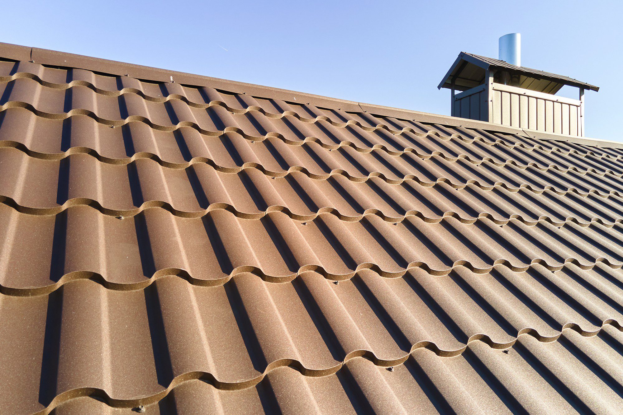 The image shows a close-up view of a brown corrugated metal roof. You can see the wavy pattern which is characteristic of this type of roofing material. The ridges add strength and allow for water runoff. Towards the top right corner of the image, there's a structure that appears to be a chimney or vent stack protruding from the roof, complete with a small protective cap or housing at the top. The sky is clear and blue, indicating a day with good weather. No people are visible in the image.