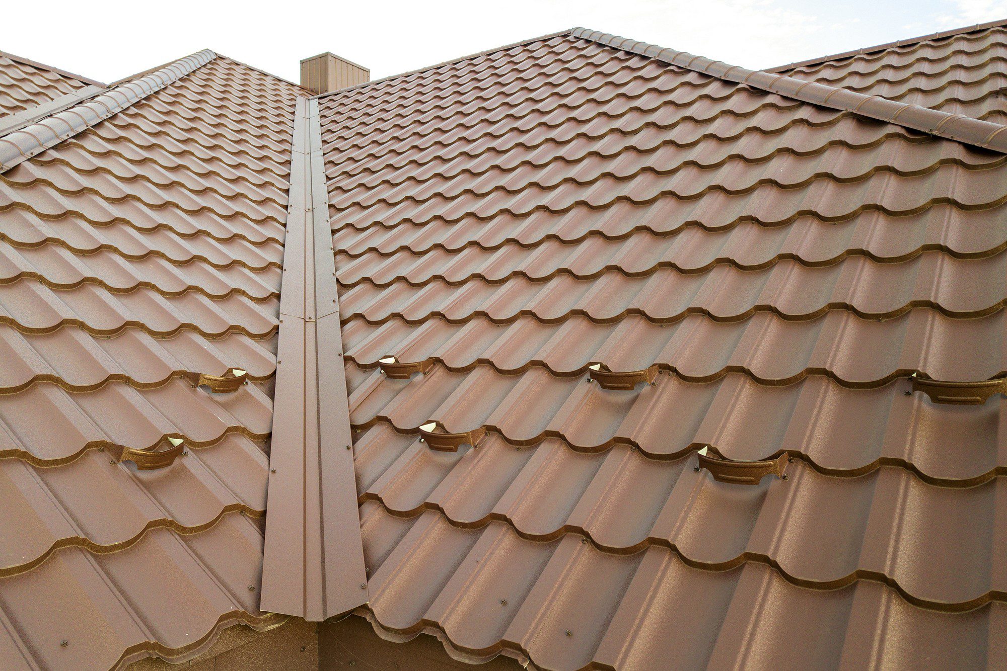 This image shows a close-up view of a tiled roof. The tiles are arranged in a repeating pattern, and they appear to be terracotta in colour, which is common for clay or ceramic roofing materials. There are also some metal elements affixed to the tiles which are likely to be snow guards or similar devices aimed at preventing snow from sliding off the roof all at once. The roof has multiple sloped sections, meeting at raised ridges, which are typical features designed for efficient water runoff. On the ridge, there is additional metal flashing, likely for waterproofing and protection where the roof planes intersect.