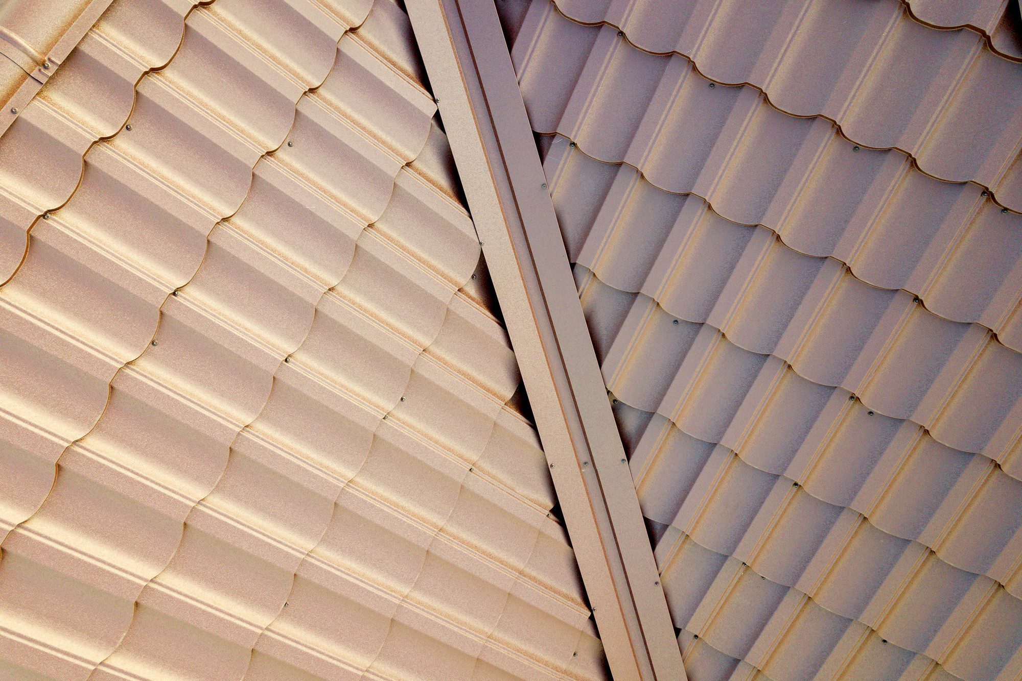 This image shows a close-up view of metal roofing material. It features corrugated metal sheets, characterized by their regular, undulating pattern of ridges and grooves which add strength and rigidity to the material. The sheets are fastened to a structure using screws, as seen periodically along the ridges. The brownish tone of the sheets suggests either a paint finish or a natural rust-resistant coating, such as those used for weatherproofing and aesthetic purposes. The overlapping sections and the linear nature of the joints between the sheets ensure water runoff and contribute to the roofing's waterproof properties. The angle and composition of the photo create an interesting pattern with geometric appeal.