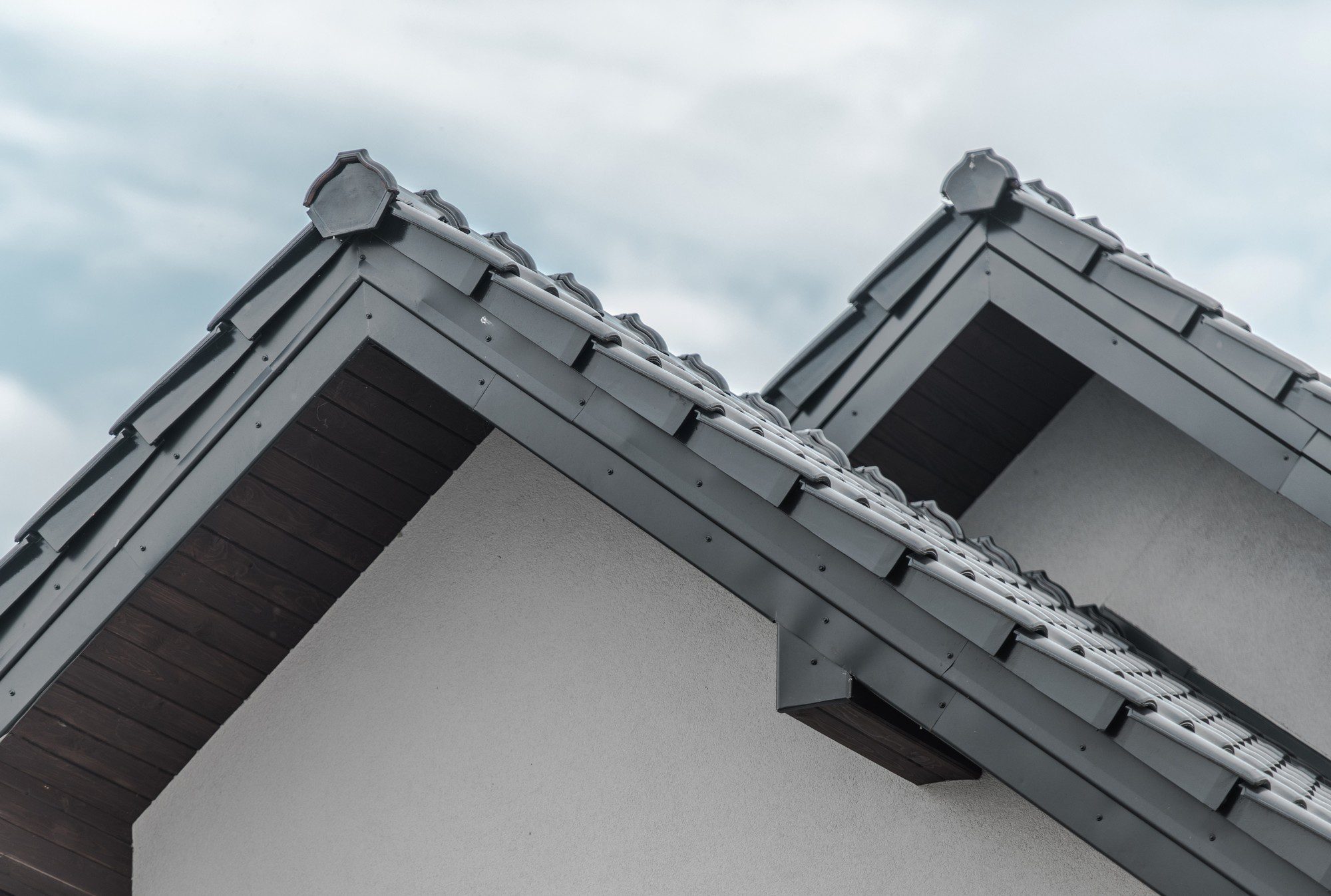 This image shows the upper part of a building focusing on a detail of the roof. The roof is covered with gray tiles and features two ridgelines with ridge caps. There is a gutter system at the edge of the roof to handle rainwater. The siding under the roof appears to be wood or a wood-like material. This architectural detail is typical for pitched roofs designed to promote water runoff and prevent water infiltration into the building. The sky in the background is overcast.