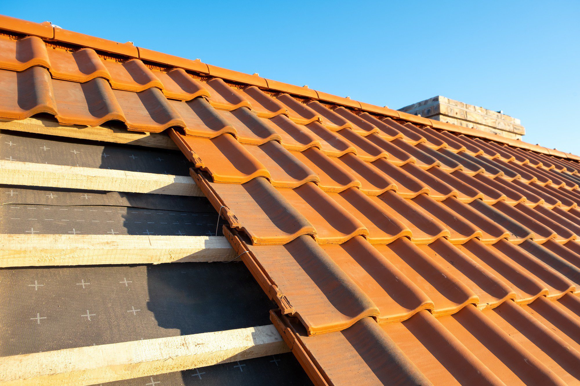 This image shows a close-up view of a terracotta tiled roof. The roof tiles have a wavy pattern, typical for clay or ceramic tiles used in many regions for roofing. There is a clear blue sky in the background, suggesting it might be a sunny day. The photo captures the roof at an angle that highlights the overlapping pattern of the tiles and also shows the roofing underlayment beneath the partially completed section of the roof. The underlayment appears to be a black material, likely waterproof, which provides an additional layer of protection against the elements. Wooden battens can be seen where the underlayment is exposed; these battens are used to anchor the tiles in place.