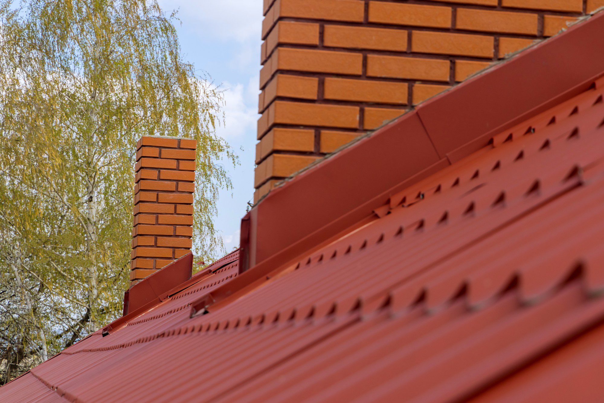 This image shows a section of a building's exterior, focusing on a red metal roof with a pattern of raised ridges and exposed fasteners. The roof has a metallic sheen, indicating its metallic construction, likely steel or aluminium with a red coating for weather resistance and aesthetic purposes. On the left side, there is a brick chimney extending upwards, with a clear sky behind it and the branches of a leafy, possibly birch, tree partially visible, suggesting a residential setting. The perspective is slightly angled upwards from the roof towards the chimney, creating a sense of depth.