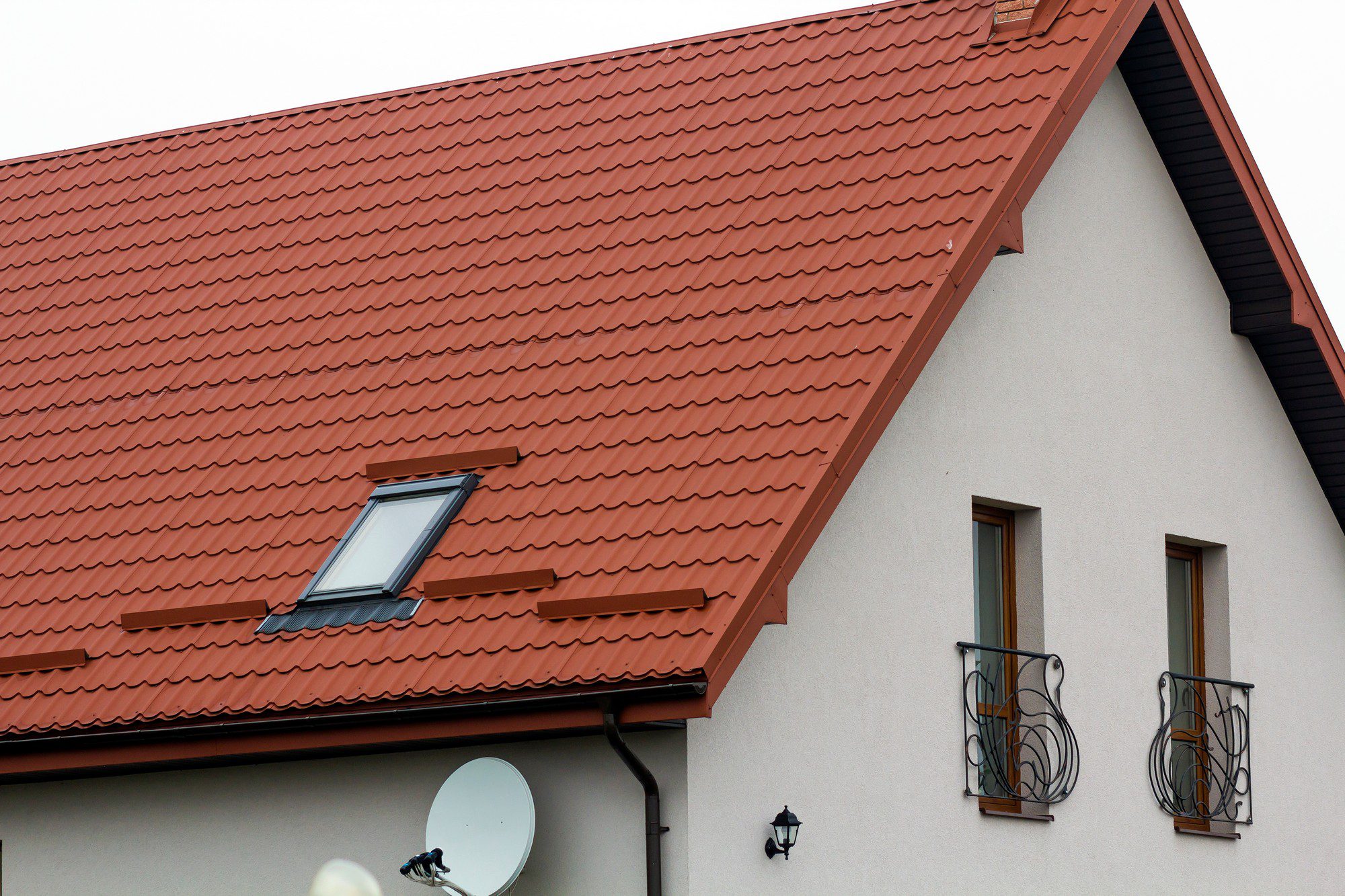 The image shows a section of a building featuring a gable roof covered with terracotta-coloured roof tiles. There is a single dormer window on the roof with a small roof section of its own. The side wall of the house is painted in a light colour and has two windows; both windows have decorative metal grilles and shutters on the outside. Also visible on the wall are a small outdoor wall lantern and a satellite dish with a bird perched on top of it. The overall style suggests a modern residential building with some traditional design elements.