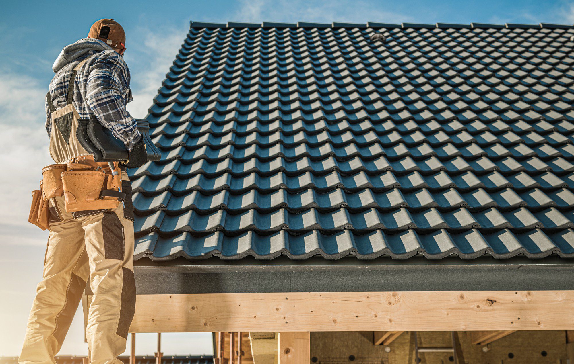 This image features a construction worker on a rooftop. The worker is wearing a tool belt and is seen from behind. They are looking at a dark-coloured tiled roof that appears to be newly installed or in the process of being worked on. It is a sunny day with a clear blue sky in the background, and the worker is standing on a wooden structure that supports the roof, which seems to be in the process of being constructed or repaired.