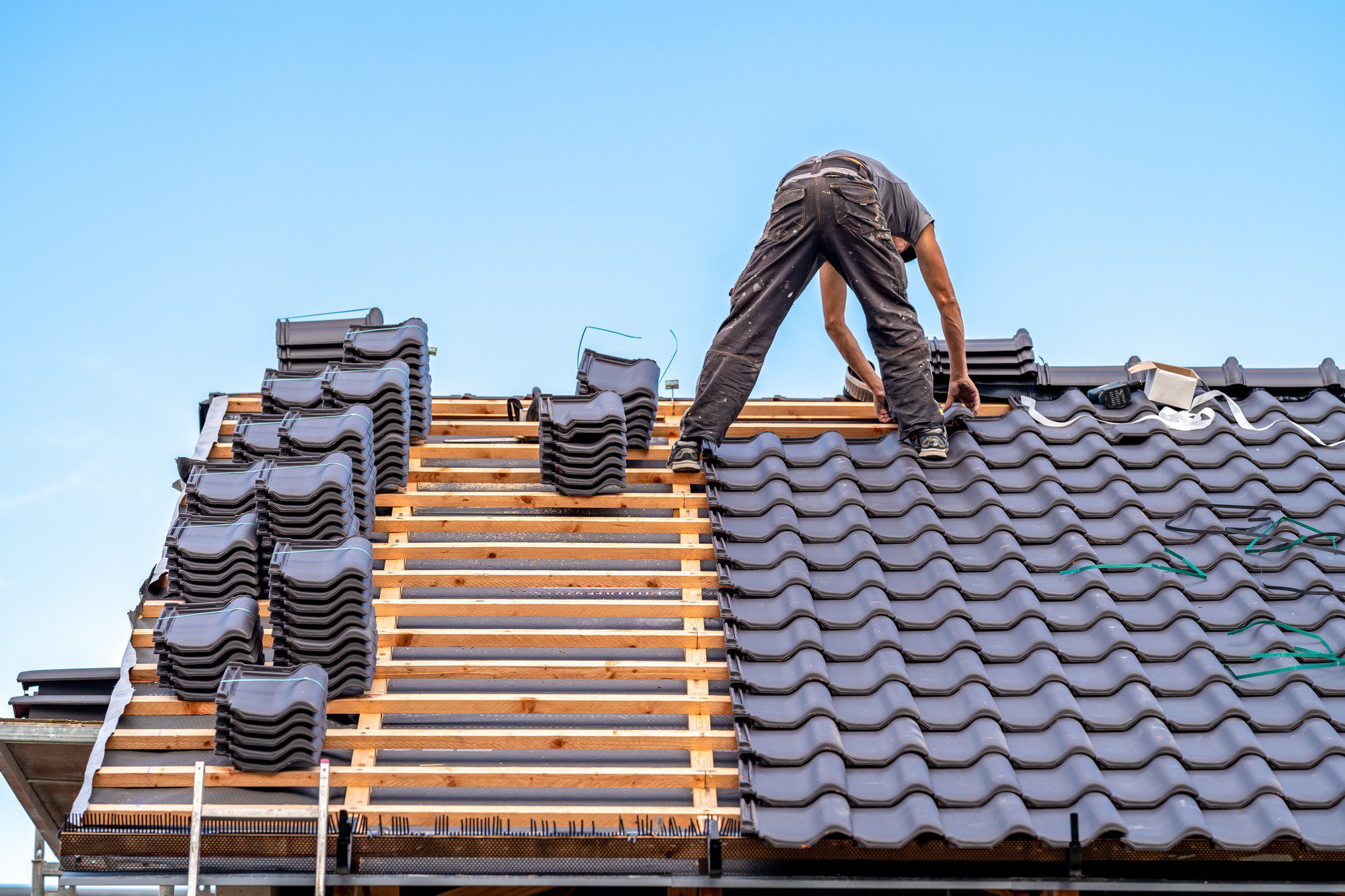 This image depicts a person working on a roof. They appear to be installing or repairing roofing tiles. The person is wearing work clothes and is bending over as they handle the roofing materials. The roof structure is partially complete, with some areas covered by black tiles and others exposing the wooden slats underneath. The stacks of tiles are arranged on the roof, ready for installation. The sky is clear, suggesting good weather for outdoor construction work.