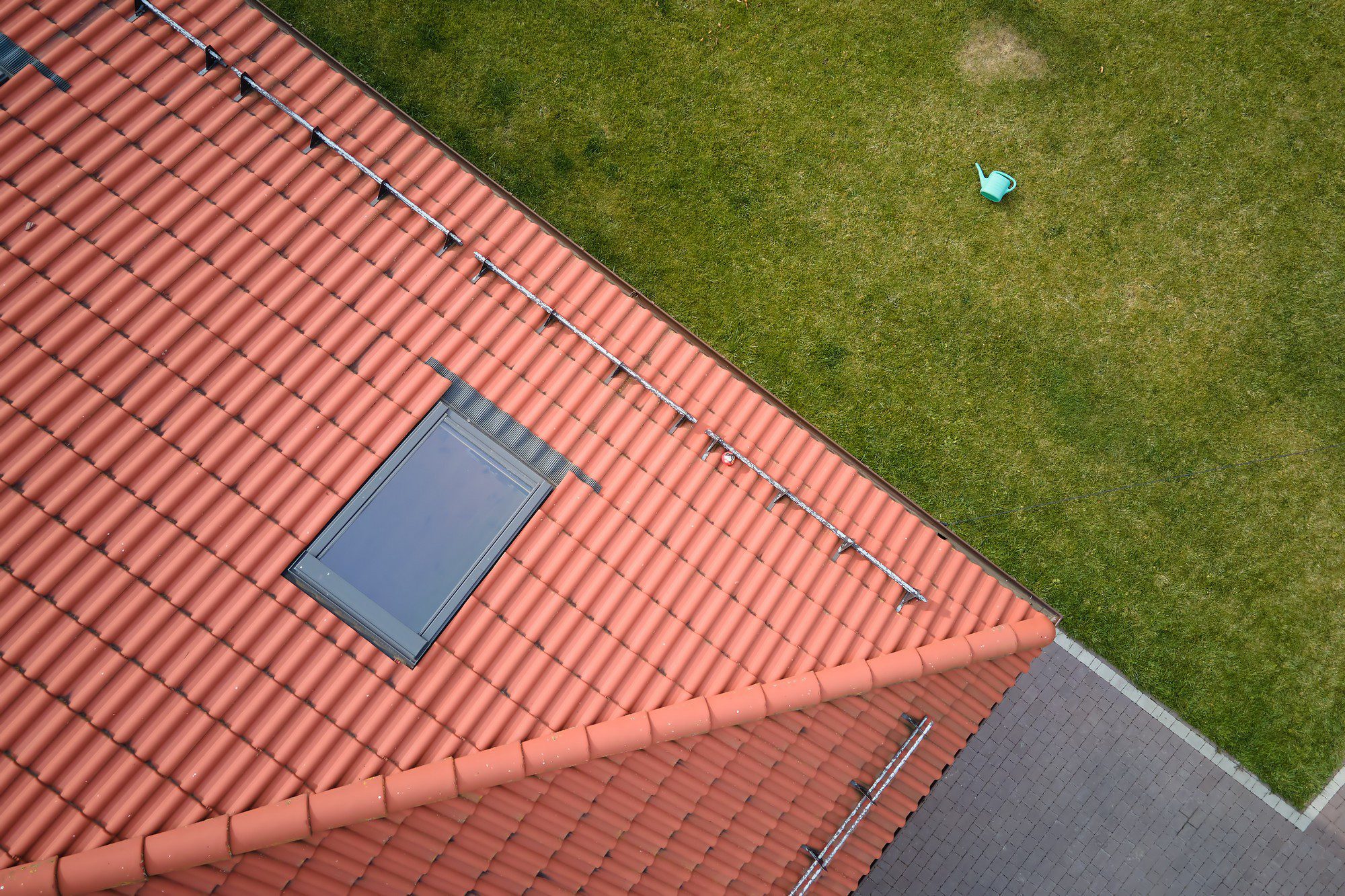 This image features a high-angle view of part of a building with a terracotta-tiled roof. There's a skylight window on the roof. To the right of the roof, there's a well-manicured green lawn and a small blue watering can lying on the grass. In the bottom right corner of the image, you can see what appears to be a paved area or walkway with rectangular gray paving stones. The perspective suggests the photo was taken from an upper floor of a nearby building or structure, giving a bird's-eye view of the scene.