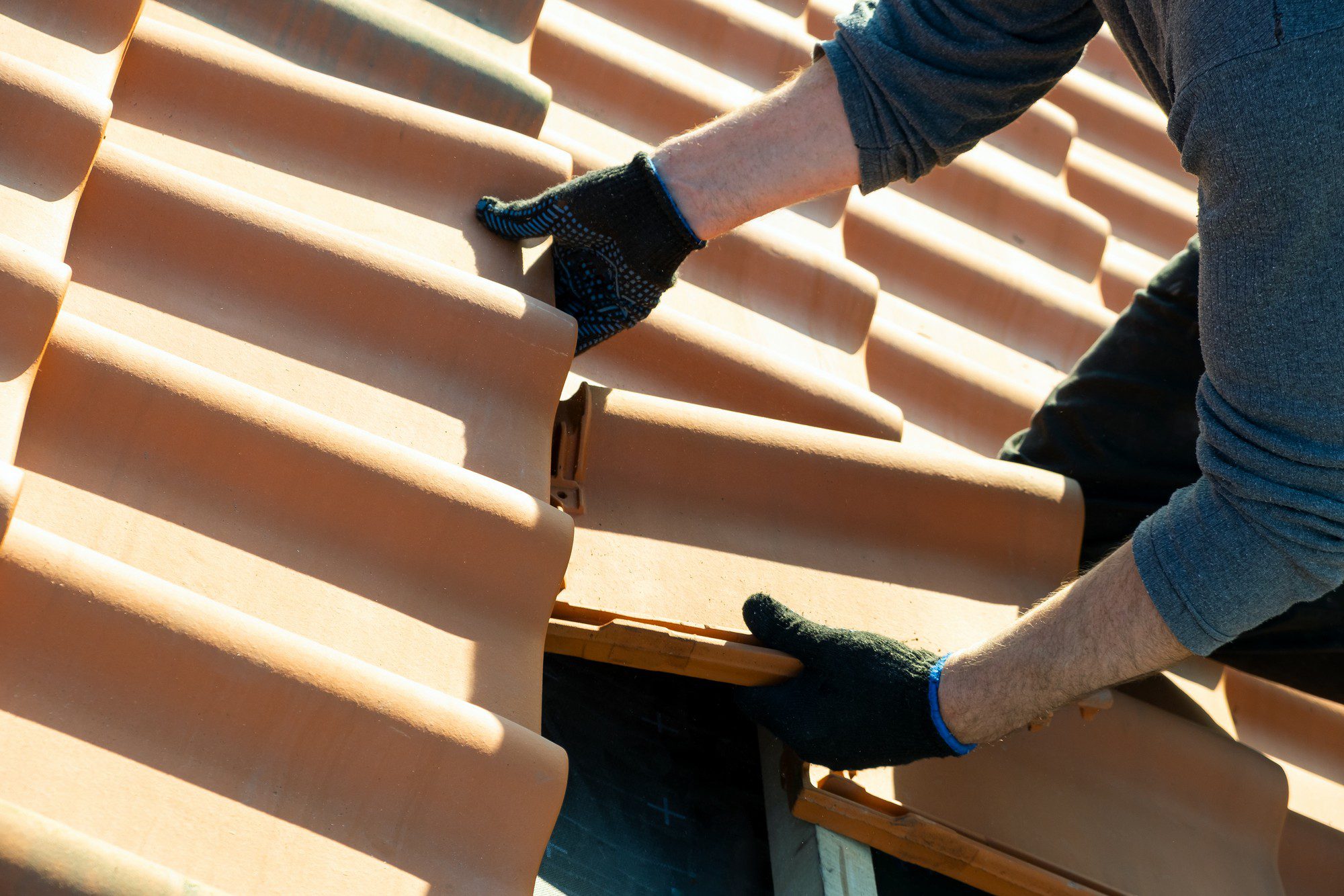 The image shows a close-up of a person's hands wearing gloves and working with terracotta roof tiles. These tiles are typically used for roofing and are known for their durability and ability to withstand various weather conditions. The person appears to be installing or repairing a roof, as they are handling the tiles carefully to put them in place or adjust them. The sunlight suggests that the work is being done outdoors during the day.