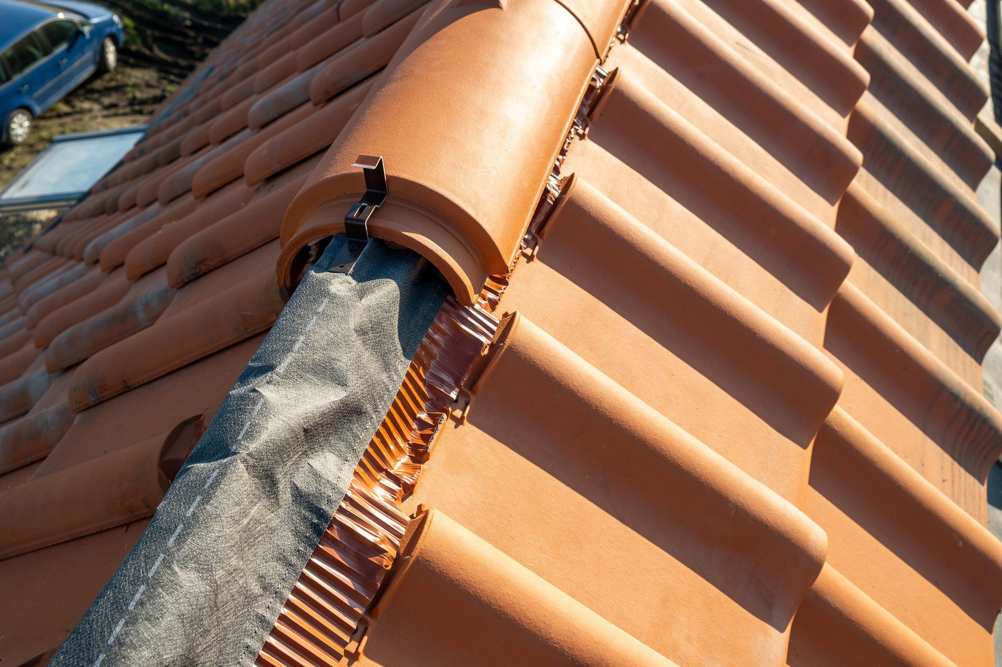 The image shows a close-up view of a section of a tiled roof. The roof tiles appear to be terracotta in colour and are the rounded, barrel-style type commonly seen in Mediterranean or Spanish-style architecture. There is a ridge vent covered with a protective fabric-like material, likely for ventilation purposes while preventing debris and pests from entering. The ridge vent, usually placed along the apex of the roof, allows for air circulation under the roof covering, which is important for temperature and moisture control inside the building. The background includes a view of the sky and the corner of a car, indicating that the photo was taken outside during a day with clear weather.