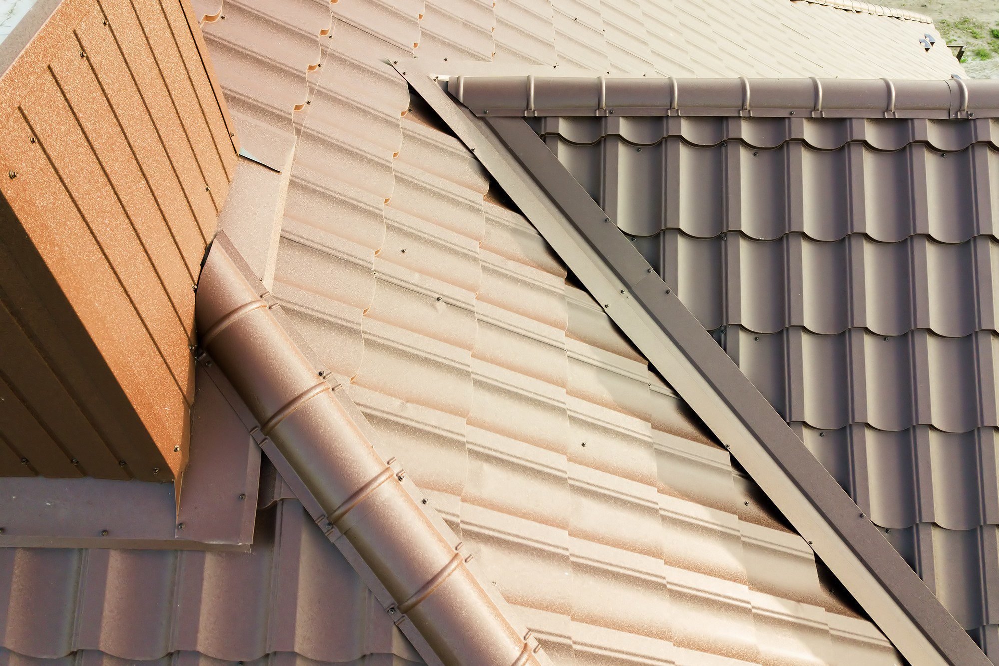 The image shows a close-up view of the interconnection between two sections of a roofing system. There are two different colours and styles of roof tiles: on the left, there are reddish-brown terracotta style tiles, while on the right, there are grey metal tiles with a corrugated pattern. A gutter downspout, matching the reddish-brown tiles in colour, runs along the intersection where the two different roofs meet, indicating an effort to manage rainwater runoff efficiently. The metal flashing along the seam where the two roofs meet helps to ensure waterproofing at the junction. The sunlight and shadows on the roof suggest a bright, sunny day.