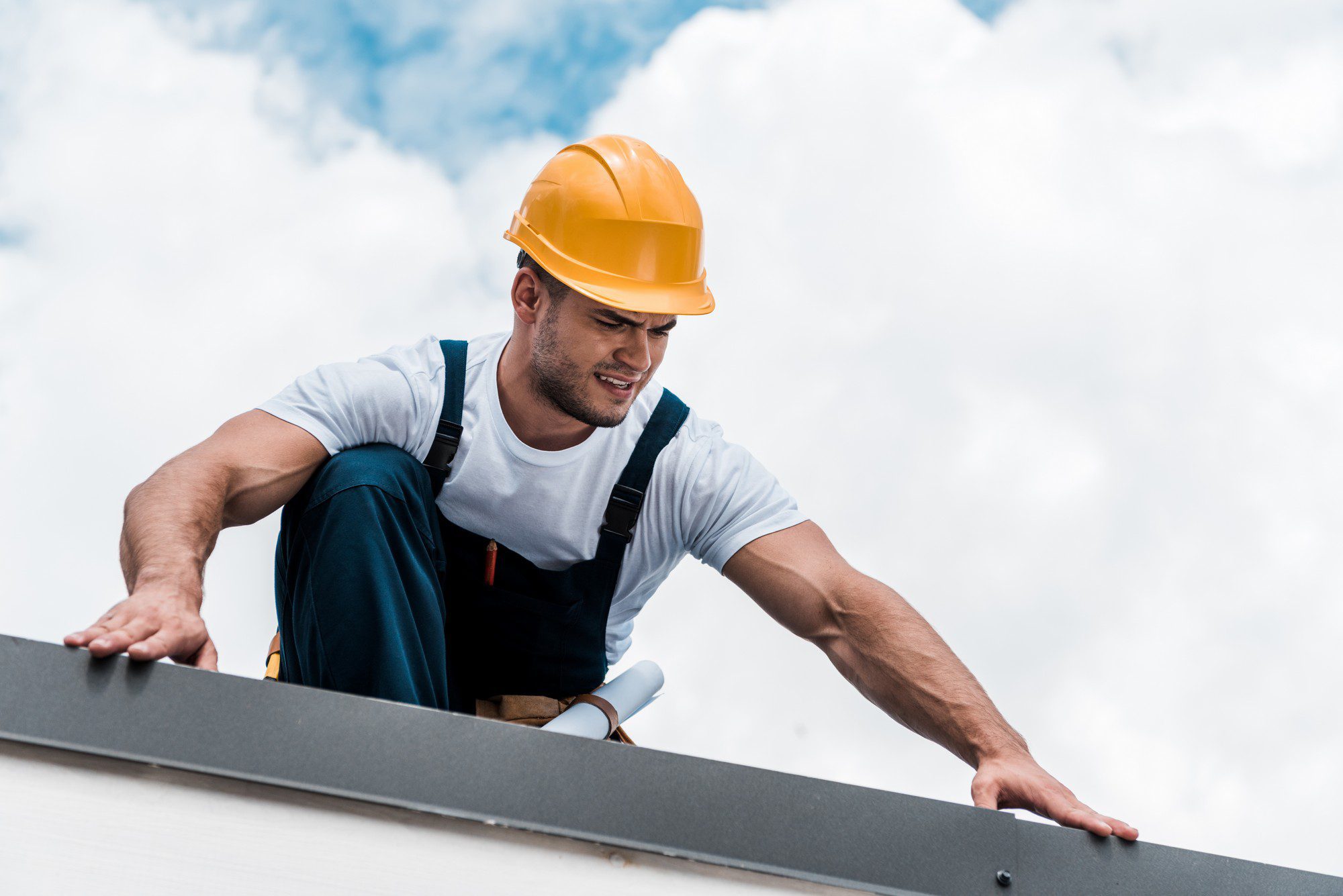 The image depicts a construction worker who appears to be either working on a high structure or inspecting it. He is wearing a safety helmet, indicative of standard safety practices on construction sites. The worker also has a tool belt, suggesting he may be performing some type of manual labour or repair work. The background shows a clear sky with clouds, indicating that the picture is taken outdoors during the daytime.