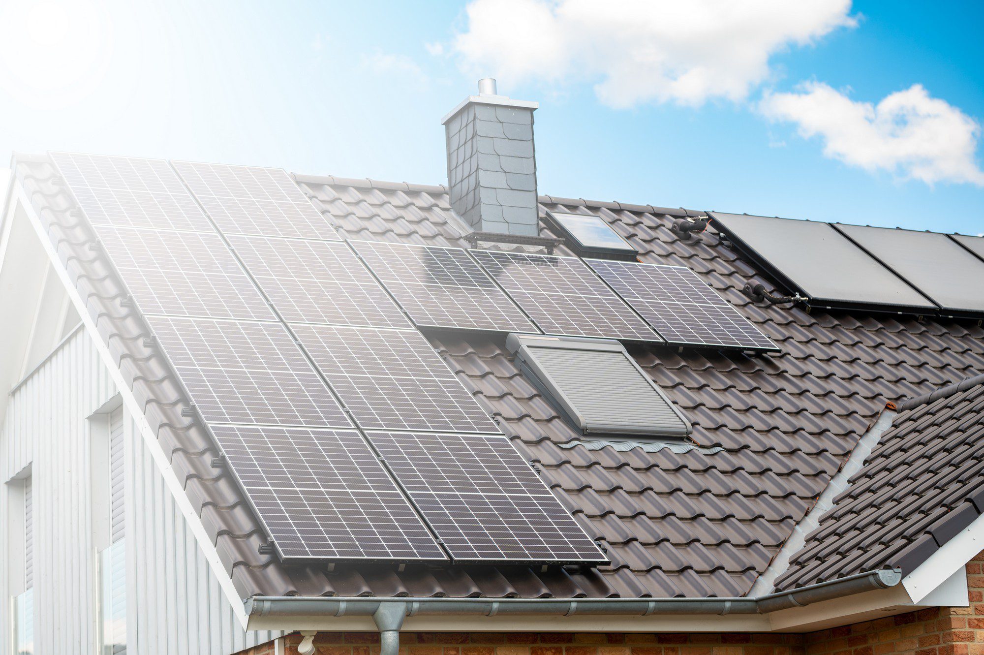 This image depicts a residential rooftop outfitted with solar panels. The solar panels are installed to harness solar energy, converting it into electricity for use in the home, thereby promoting renewable energy and potentially reducing electricity bills. The roof has a pitched design and is covered with interlocking tiles, characteristic of many suburban homes. There's also a brick chimney visible, and the sky indicates it's a bright, sunny day, which is ideal for generating solar power. The guttering system along the edge of the roof is also visible.