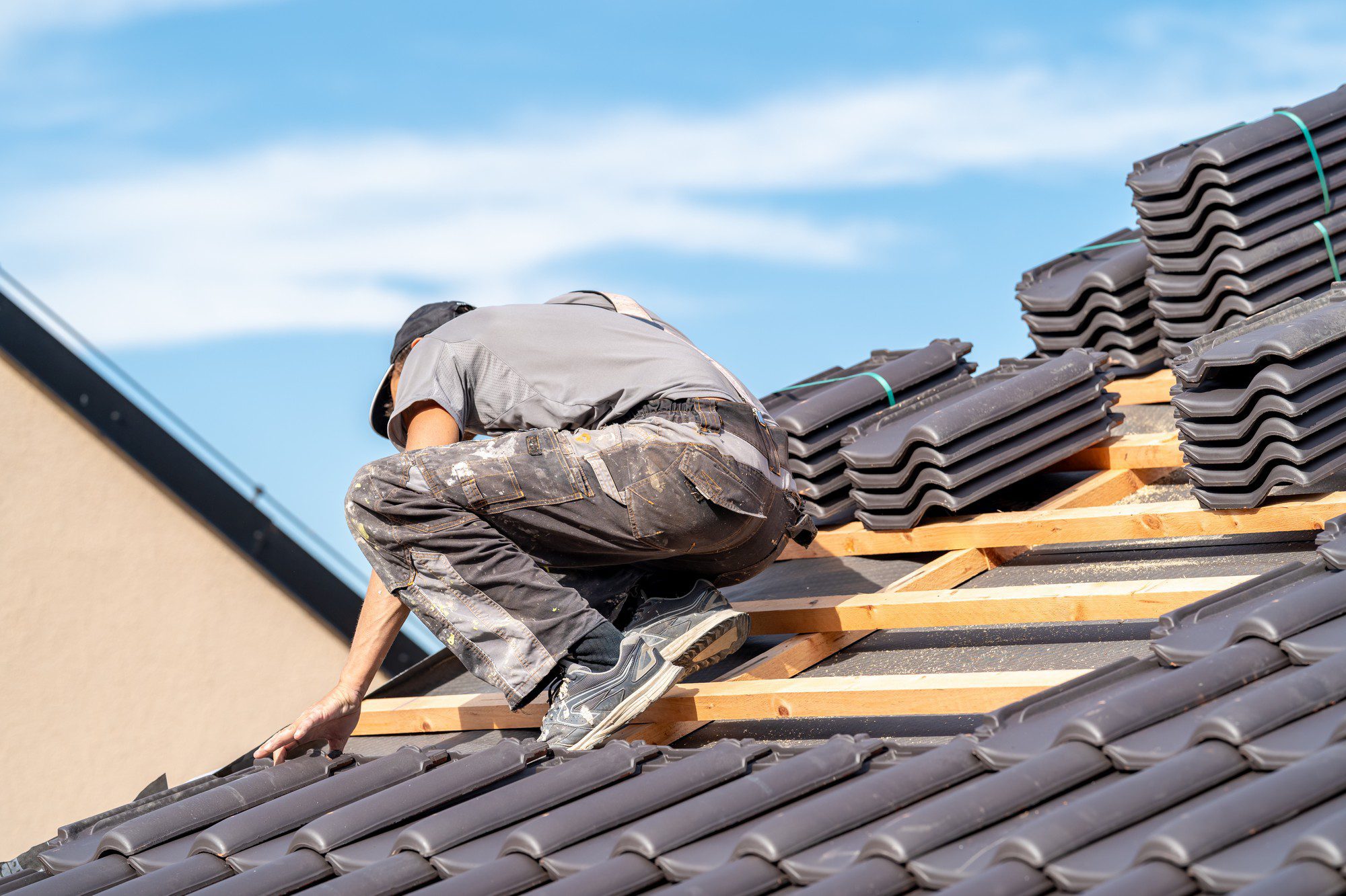 In this image, there is a person working on the roof of a building. They are on a steeply pitched roof that is partially covered with roofing tiles. The person appears to be wearing work clothing and safety equipment, suggesting they are a construction worker or roofer. There are stacks of additional tiles on the roof, indicating that the roofing process is underway. The sky is clear with a few clouds, suggesting good weather conditions for outdoor construction work. The person is bending over, likely installing or adjusting the tiles. Safety measures, like a secure footing and proper safety gear, are important when working at such heights to prevent accidents.