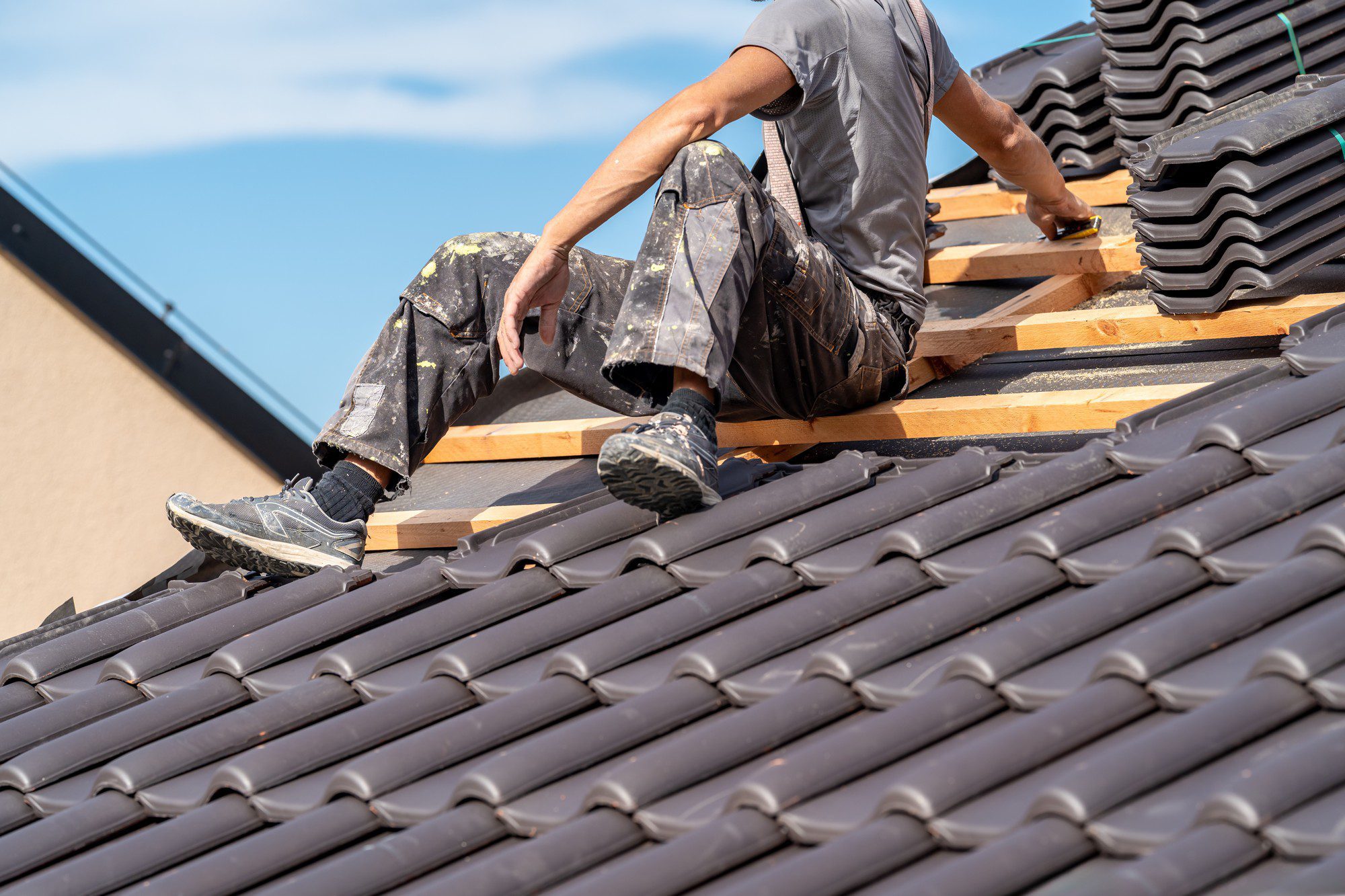 The image shows the lower half of an individual sitting on a sloped roof, working on roofing construction. This person is wearing stained work pants and trainers, indicative of manual labour, and is holding a tool – possibly a tape measure – in their right hand. Wooden battens are visible on the section of the roof where the tiles have not yet been laid, and the person appears to be in the process of installing or repairing a tiled roof. The sky is clear, suggesting fair weather conditions which are suitable for outdoor construction work.