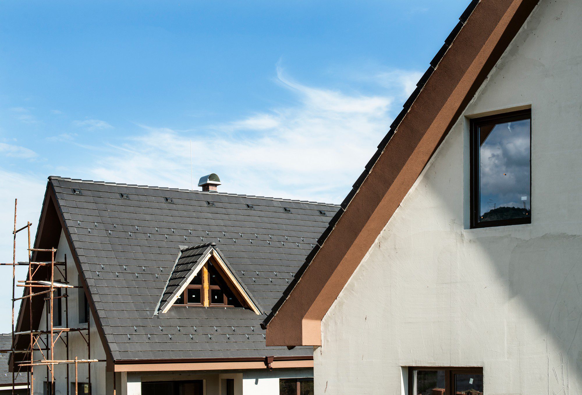 This image shows a part of a residential building under construction or renovation. On the left, there's a pitched roof with dark shingles and a dormer window, also with a pitched roof, providing an opening for natural light to enter the upper floor. One can notice a chimney rising above the ridge of the roof and scaffolding set up alongside the structure, indicating ongoing work.

On the right, we see a section of the building's exterior wall with a large window reflecting the blue sky and some clouds. The facade is finished with a white render, while the gable and window frames are accented in brown. The image captures a clear day with favorable weather for construction activities.