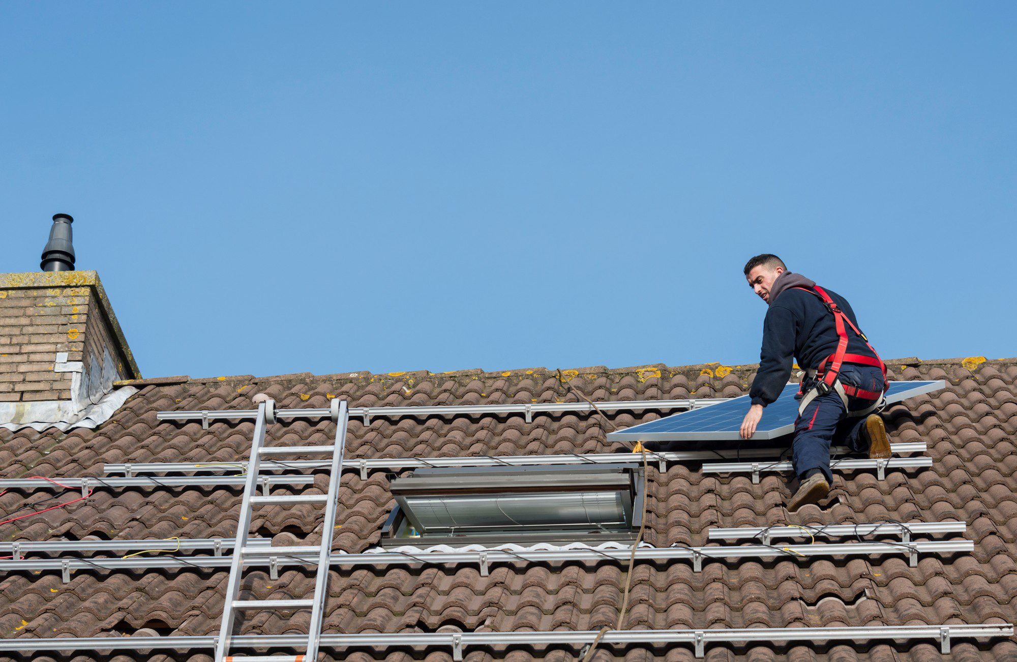 The image shows a person on the roof of a house. The person appears to be a worker, likely involved in roofing or installing solar panels, as evidenced by the solar panels already present on the roof. The worker is wearing a safety harness, which is a standard safety measure for individuals working at heights to prevent falls. The roof has a skylight and is covered with brown, curved tiles. On the left side of the roof, there's a chimney. Metal scaffolding is present at the edge of the roof to provide support and access for the workers during their tasks. The sky is clear, suggesting it's a day with good weather, which is typically preferred for outdoor work of this nature.