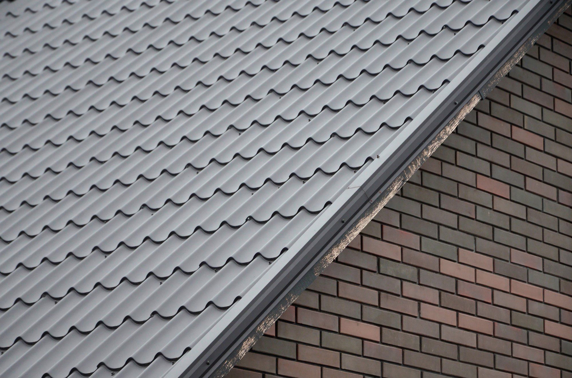 The image displays a close-up view of part of a building's exterior, showing two distinctive types of surfaces that meet at a diagonal edge. On the top section is a gray metal roof with a corrugated pattern, which is characteristic of many modern roofing materials designed for durability and effective water runoff. Below the metal roof is a section with a brick facade, made up of bricks arranged in a staggered pattern, commonly used for exterior walls due to their aesthetic appeal and structural integrity. The colour contrast between the gray metal and the reddish-brown bricks, along with the intersection of the different textures and patterns, creates a visually interesting architectural detail.