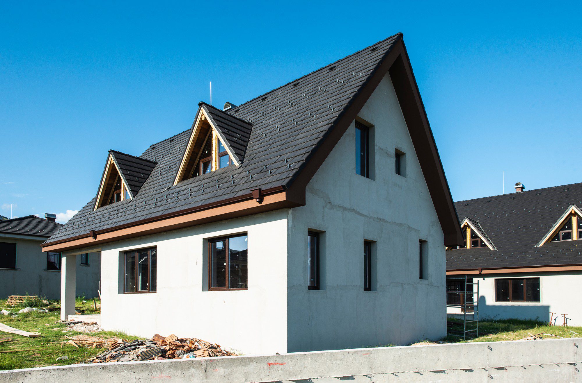 This image shows a two-story residential house under construction. The house has a pitched roof with dark shingles, and there are dormer windows projecting from the roof, which suggest additional living space or attic area upstairs. The exterior walls are finished with a light-coloured render or stucco, and the windows have not been finished yet; they are missing their final frames or exterior trim. Construction debris can be seen in the yard, indicating ongoing work. The house also features dark gutters and downspouts, and there is a scaffolding section leaning against the right side of the house, possibly for workers to complete the exterior finishing. The sky is blue, suggesting that the photo was taken on a clear day.