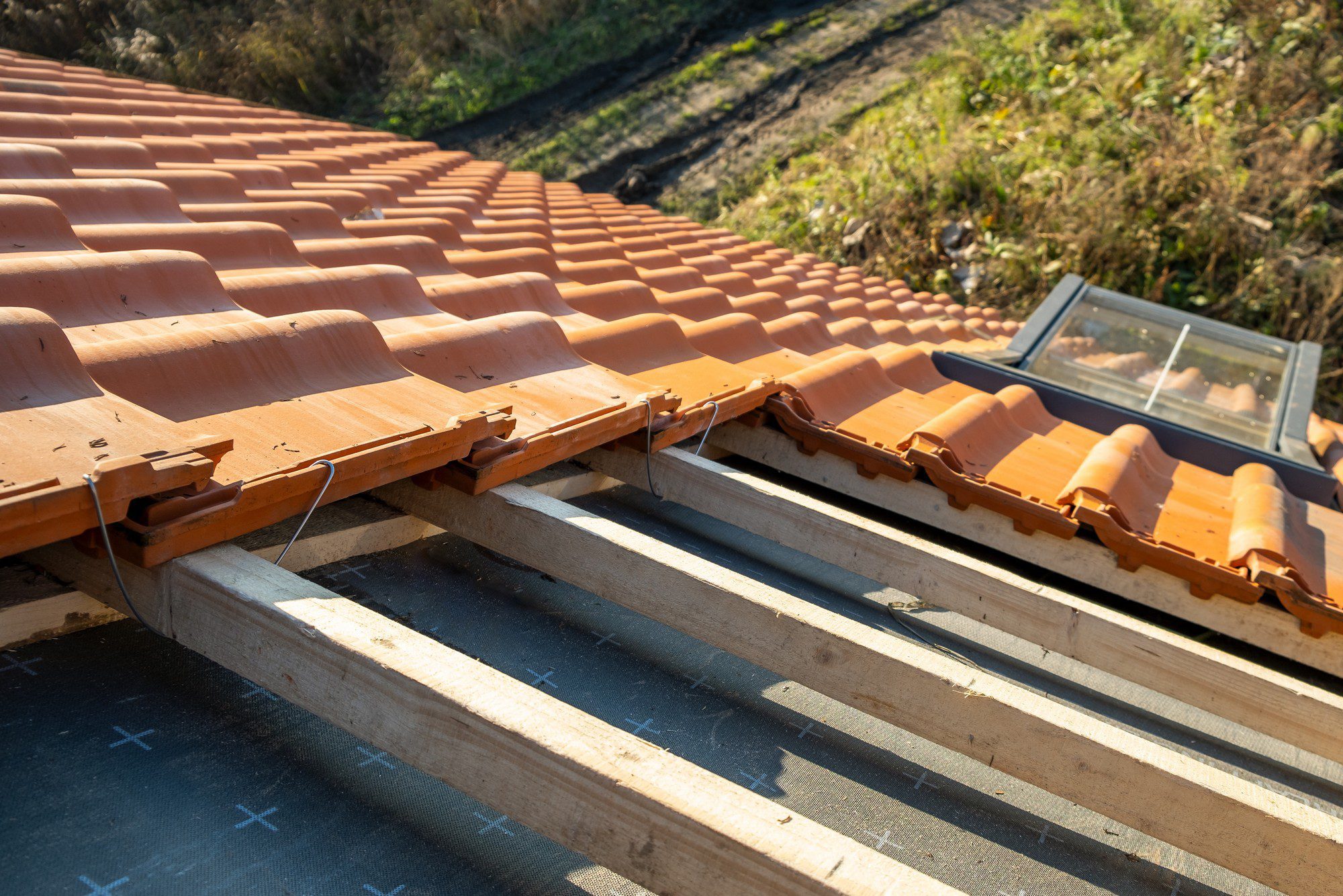 This image shows a close-up of a terracotta tile roof under construction or repair. We can see the wooden roof battens and the black roofing underlay, which is typically a water-resistant or waterproof barrier material installed directly onto the roof deck. There's also a roof window (commonly known as a skylight) installed, allowing natural light into the space below. The photograph captures the work-in-progress nature of roof installation or maintenance.