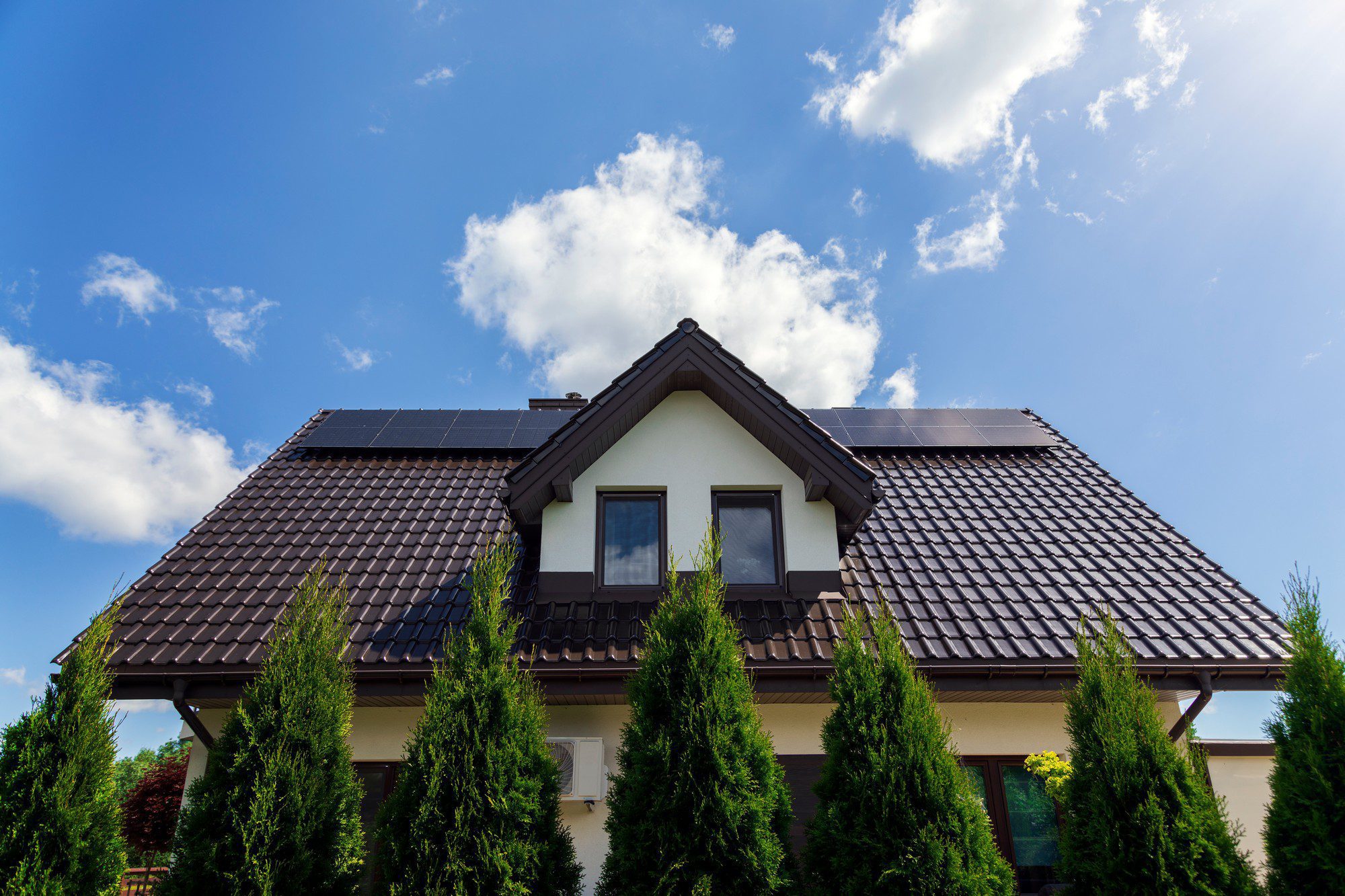 The image shows the gable end and roof of a two-story house. The roof has solar panels installed on it, which are used to convert sunlight into electricity, suggesting a commitment to renewable energy. The house has a beige façade with white trim around the windows and a dark-coloured roof. In front of the house, there is a row of tall, conical shrubs, adding greenery to the scene. The sky above is blue with some scattered clouds. This type of home improvement is becoming increasingly popular as homeowners seek sustainable energy solutions and ways to reduce their electricity bills.