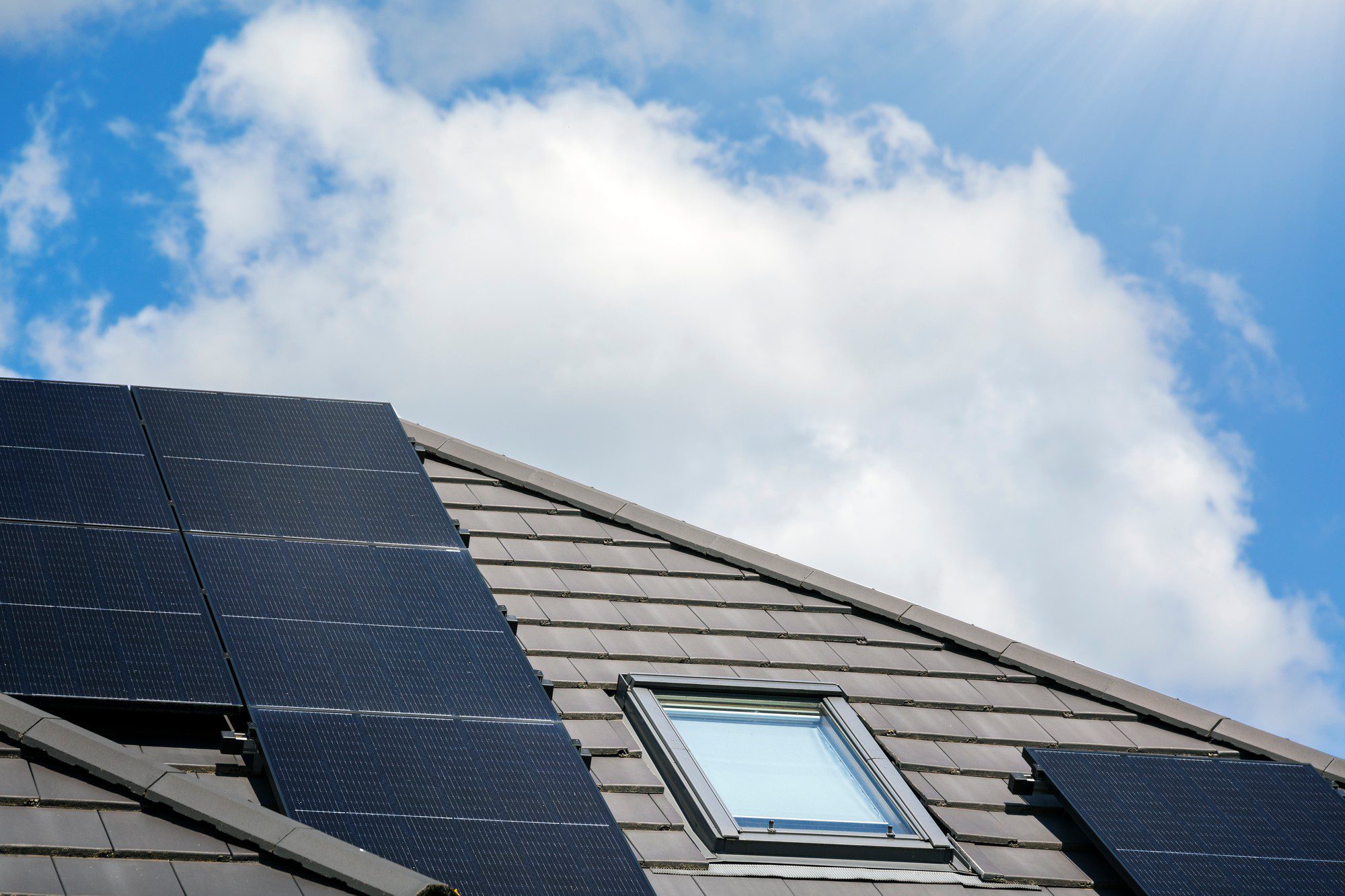 The image shows a section of a roof equipped with several solar panels, which are likely being used to generate electricity from sunlight, demonstrating the use of renewable energy in a residential setting. You can also see a skylight window installed in the slope of the roof, allowing natural light to enter the building. The sky in the background is partly cloudy with sun rays visible, indicating fair weather conditions.