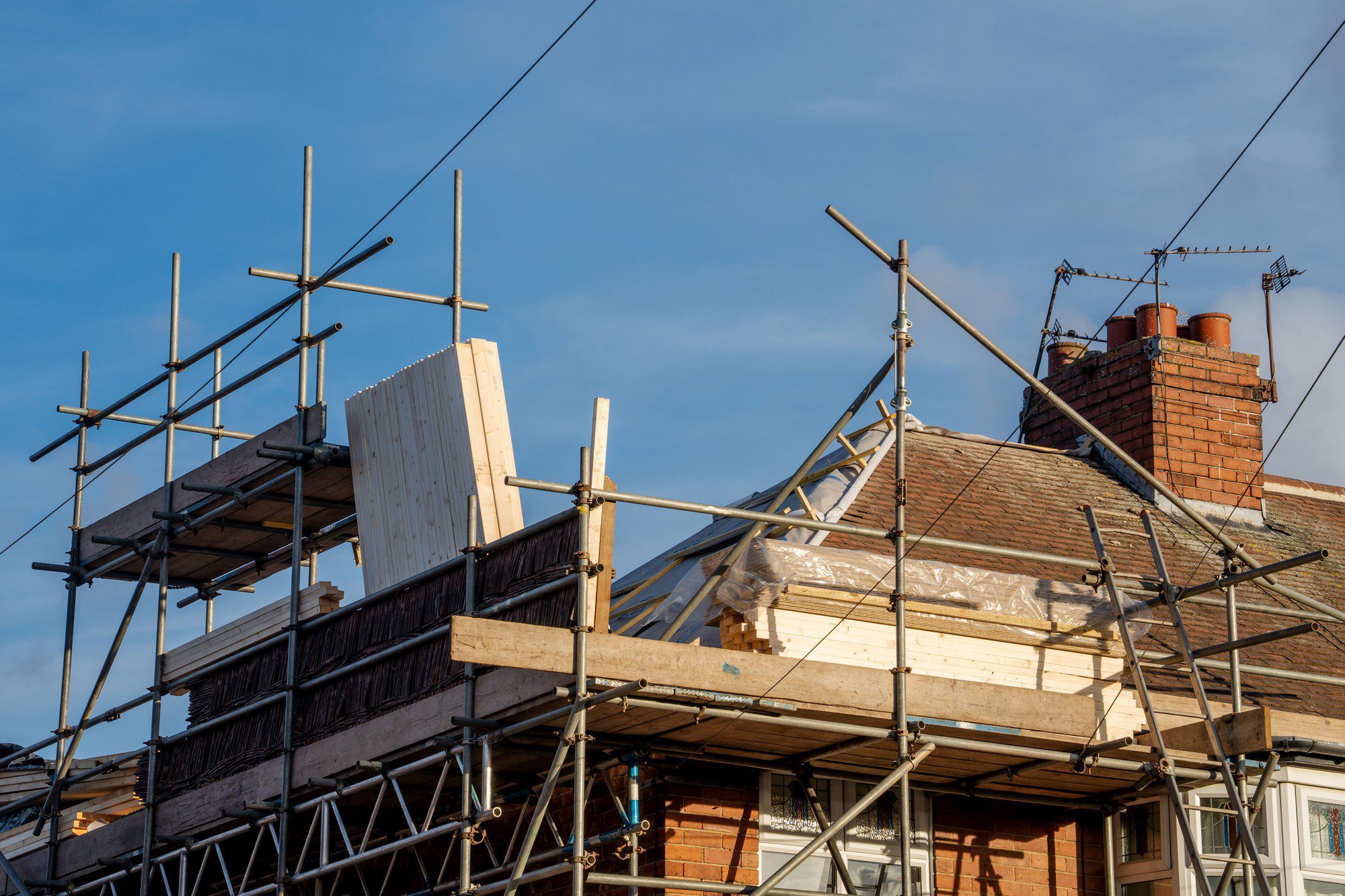 This image shows a house that is currently undergoing construction or renovation work. There is scaffolding erected around the upper part of the building, indicating that work is being done at an elevated level. Some plywood or wooden boards are visible on the top of the scaffolding platform, likely used to support workers or materials. The house has a pitched roof with roof tiles, and you can also see a brick chimney with multiple flues and a TV antenna on the top. The blue sky with some light clouds suggests it might be a fair weather day for construction work. No people are visible in the image, keeping the focus on the construction elements and the house itself.