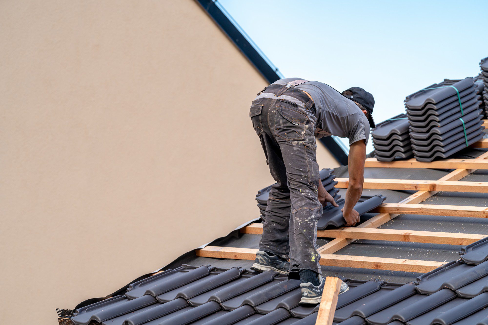 The image shows a person working on a roof. This individual is in the process of installing or repairing roofing materials, specifically tiles. The person is bent over, focusing on the task, and is wearing a cap, a pair of gloves, and work clothes which appear to be covered in dust or debris from the construction work. We can see roofing battens and underlayment, which are common materials used in roofing to provide support for the tiles and waterproofing for the structure. There is a stack of additional tiles on the roof, indicating that the work is ongoing. The sky is clear, suggesting good weather conditions for outdoor construction work.