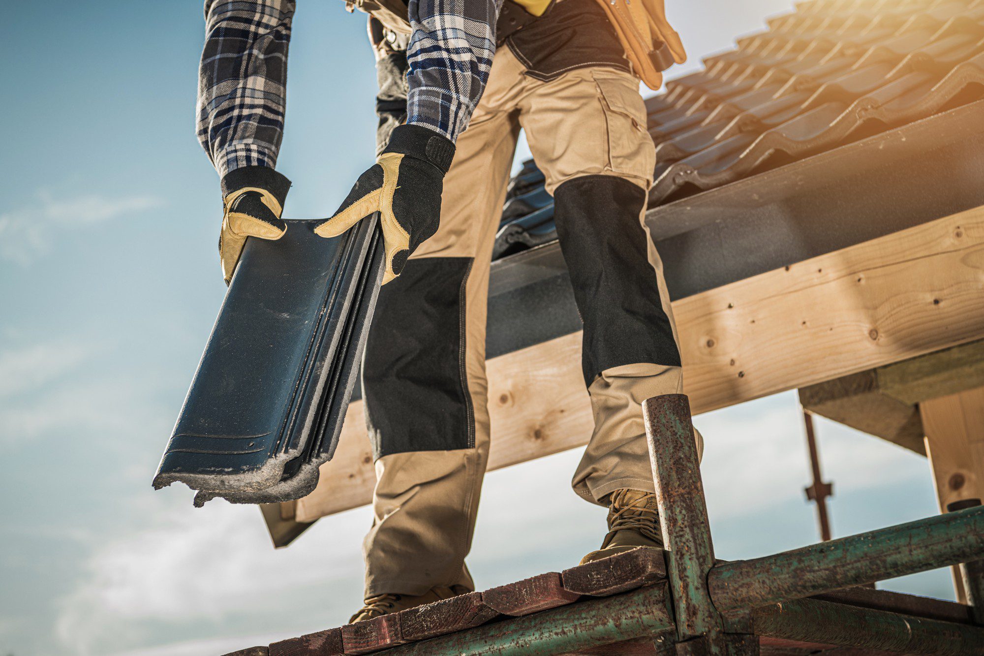 The image shows a person engaged in roofing work. The individual is standing on a scaffold and is holding a bundle of new roofing tiles. They are wearing a plaid shirt, work pants, heavy-duty gloves, and boots — typical safety attire for construction work. The background suggests a clear day with sunshine, which is ideal for outdoor construction-related activities. The focus on the hands gripping the tiles and the elevated perspective emphasizes the hands-on, physical nature of the work being performed.