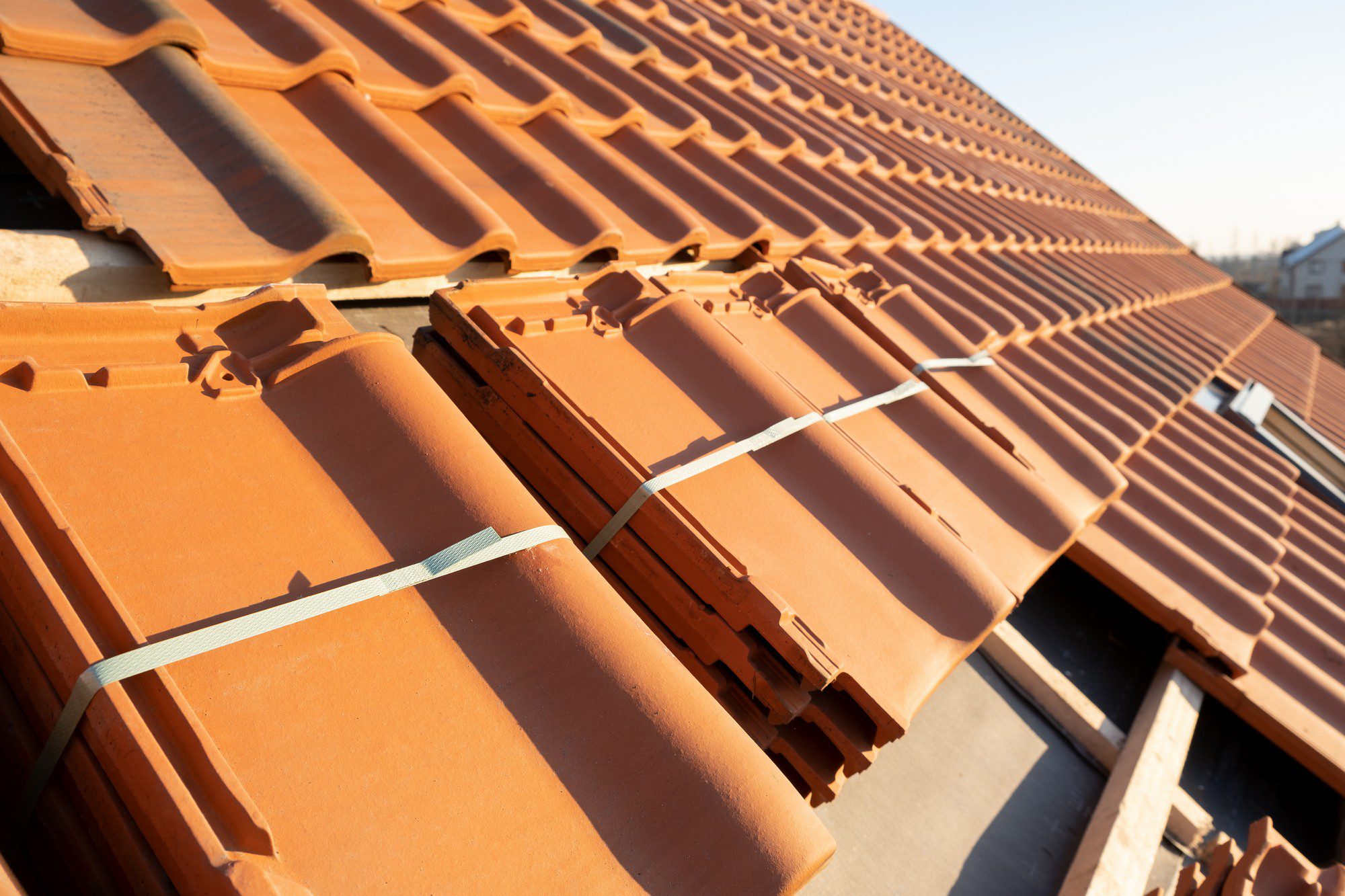 This image showcases a close-up view of terracotta roof tiles. These tiles are characterized by their wavy pattern, which is a common design for interlocking roof tiles. They are stacked and secured with metal straps, likely in preparation for installation on a roof or maybe for transportation. The perspective is such that you can see the neat rows of stacked tiles receding towards the top right of the image, and the clear blue sky suggests that it is a sunny day, casting shadows and highlighting the warm orange-red colour of the terracotta.