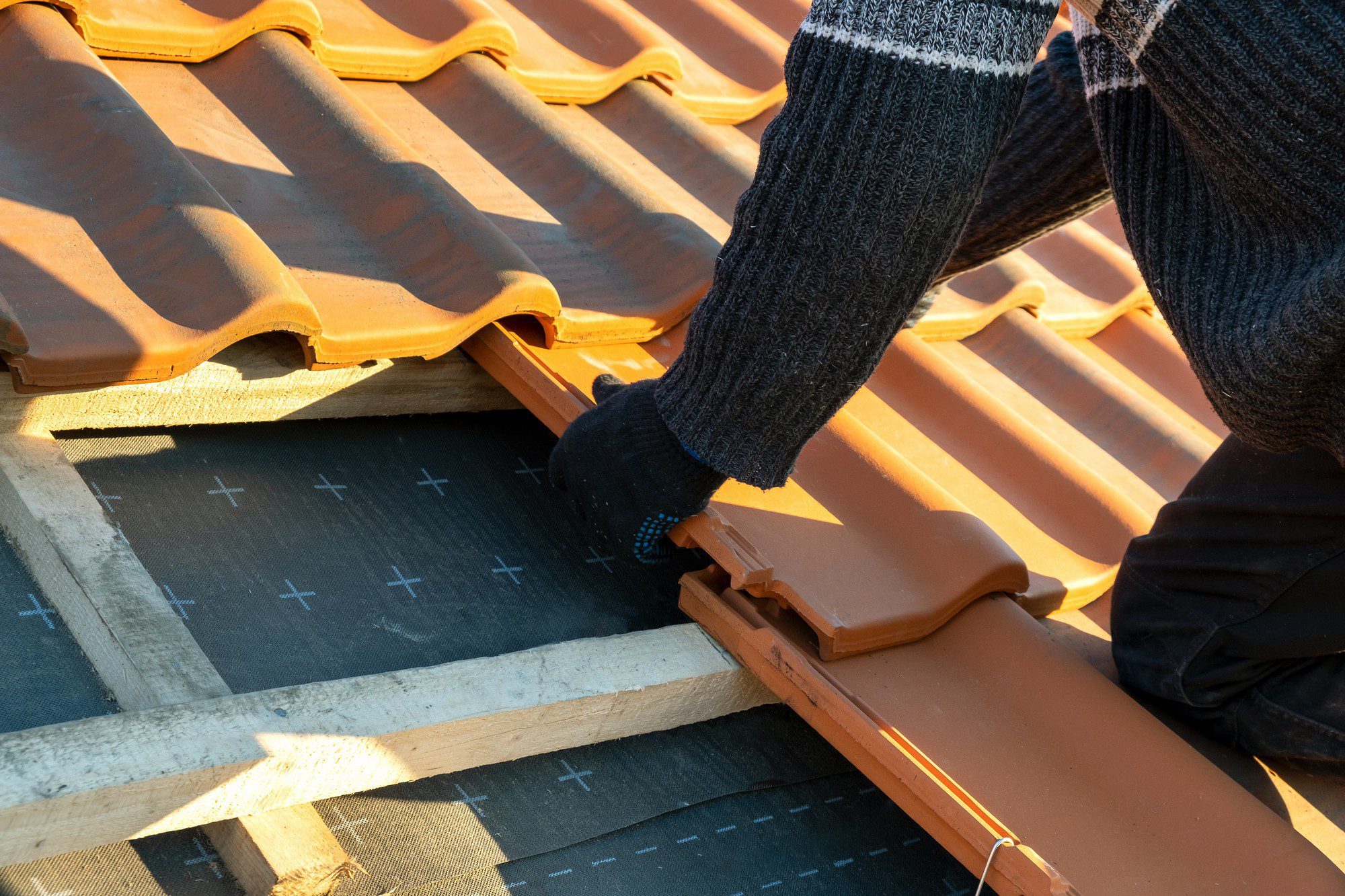 The image shows a person engaged in roofing work. Specifically, it looks like they are installing or repairing a tile roof. You can see terracotta roof tiles, some of which are lifted or being adjusted by the person. The individual is wearing gloves, indicative of manual labour, likely for protection and better grip.

Below the tiles, there is a black protective underlayer visible, which is used to provide additional waterproofing protection to the roof structure. The underlayer is laid out over wooden roof battens that create the structure to which the tiles are fixed. The person appears to be working carefully to ensure the tiles fit properly and the roof structure is secure and watertight.