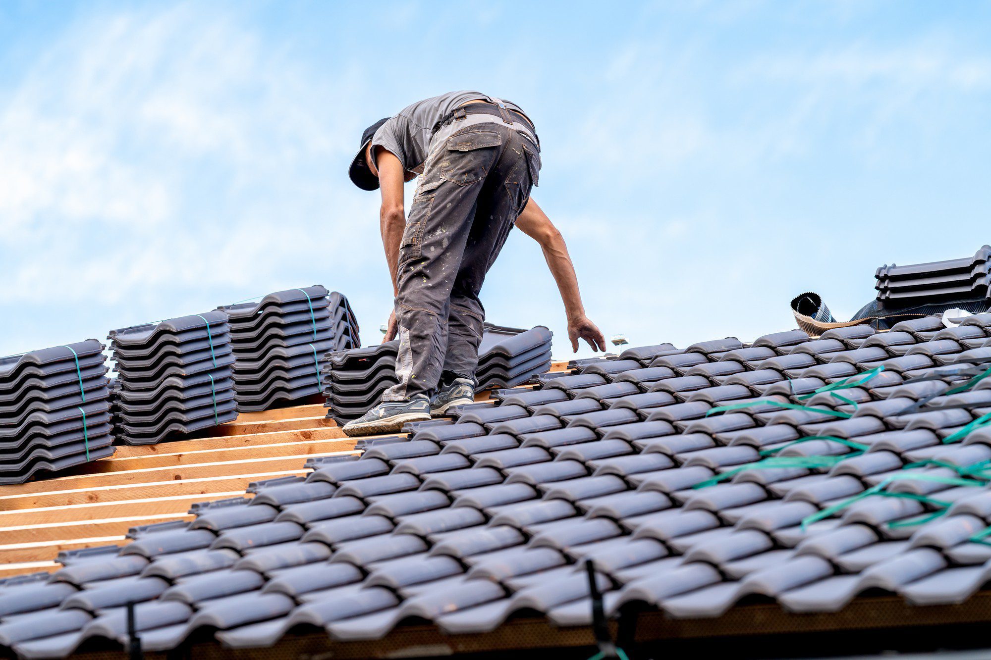 The image shows a person working on a roof. They are in the process of installing roofing tiles, which can be seen both stacked and laid out across the roof structure. The worker is bent over, handling the tiles, and appears to be engaged in manual labour, typical of roof construction or repairs. They are wearing work clothes that look appropriate for construction work, including a cap. In the background, you can see a clear sky with a few clouds. Safety measures, such as a secure footing or harnesses, cannot be directly confirmed from the image provided. The environment suggests that this could be a residential area due to the nature of the roofing work being performed.