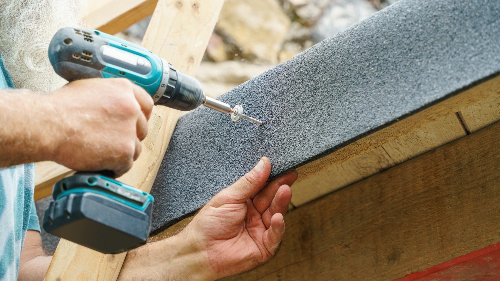 The image shows a close-up of a person using a cordless power drill to drive a screw through a piece of grey textured material into a wooden structure. The person's left hand is supporting the material as they work, and their right hand is holding the drill. The wooden structure appears to be part of some construction, possibly in an outdoor setting judging by the visible rocks in the background. The person visible has a beard, indicating they are likely male, and the focus is on the action of the hands and the drill.