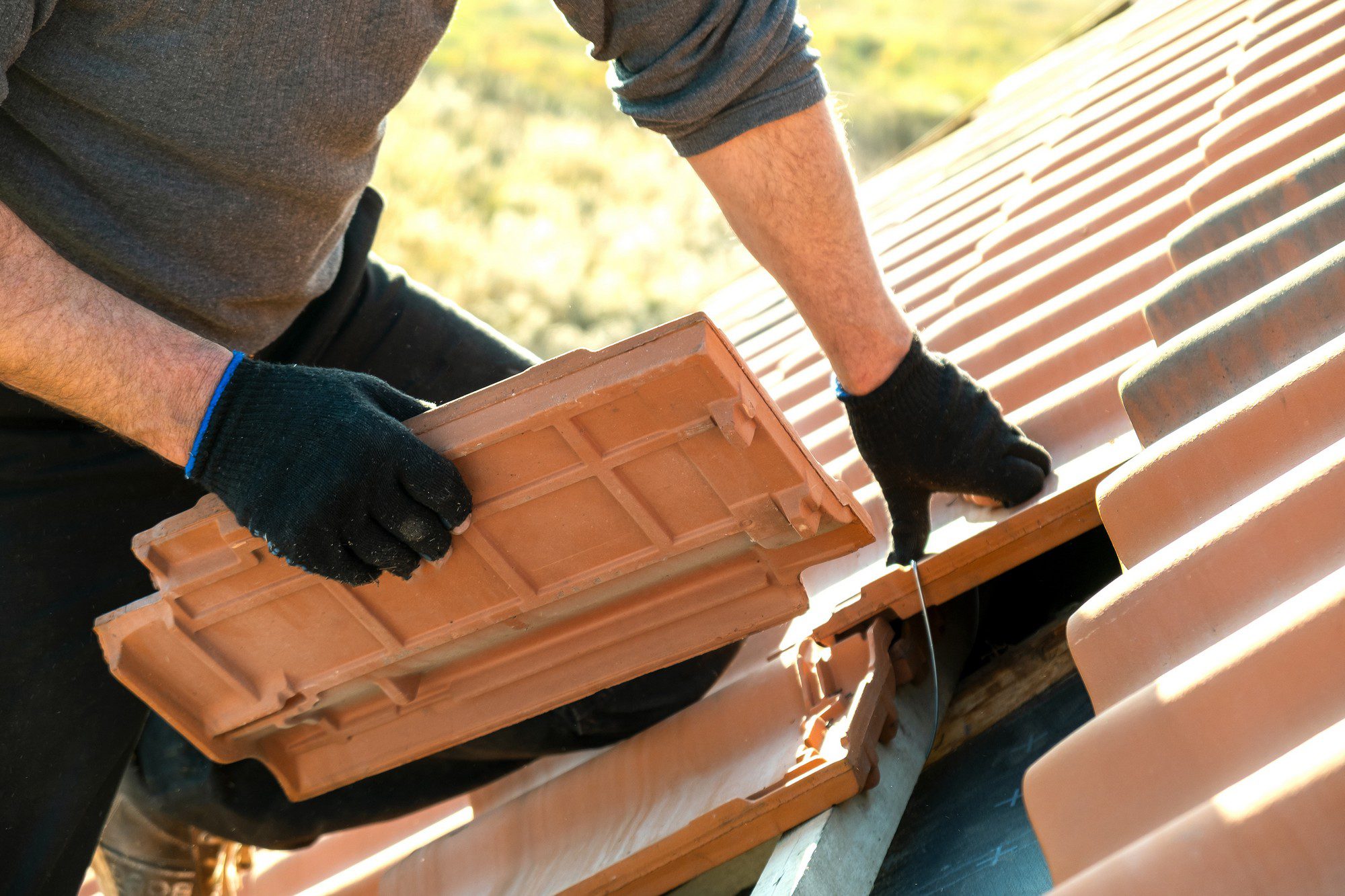 The image shows a person in the process of installing or repairing a clay tile roof. The individual is wearing gloves and has positioned a roof tile, apparently preparing to lay it into place on the roof. The picture captures hands working with the roofing material, emphasising a manual labour task. It's a sunny day, suggesting an outdoor work environment, and the angle of the shot provides a good view of the terracotta roofing tiles, which are a common material due to their durability and aesthetic appeal.