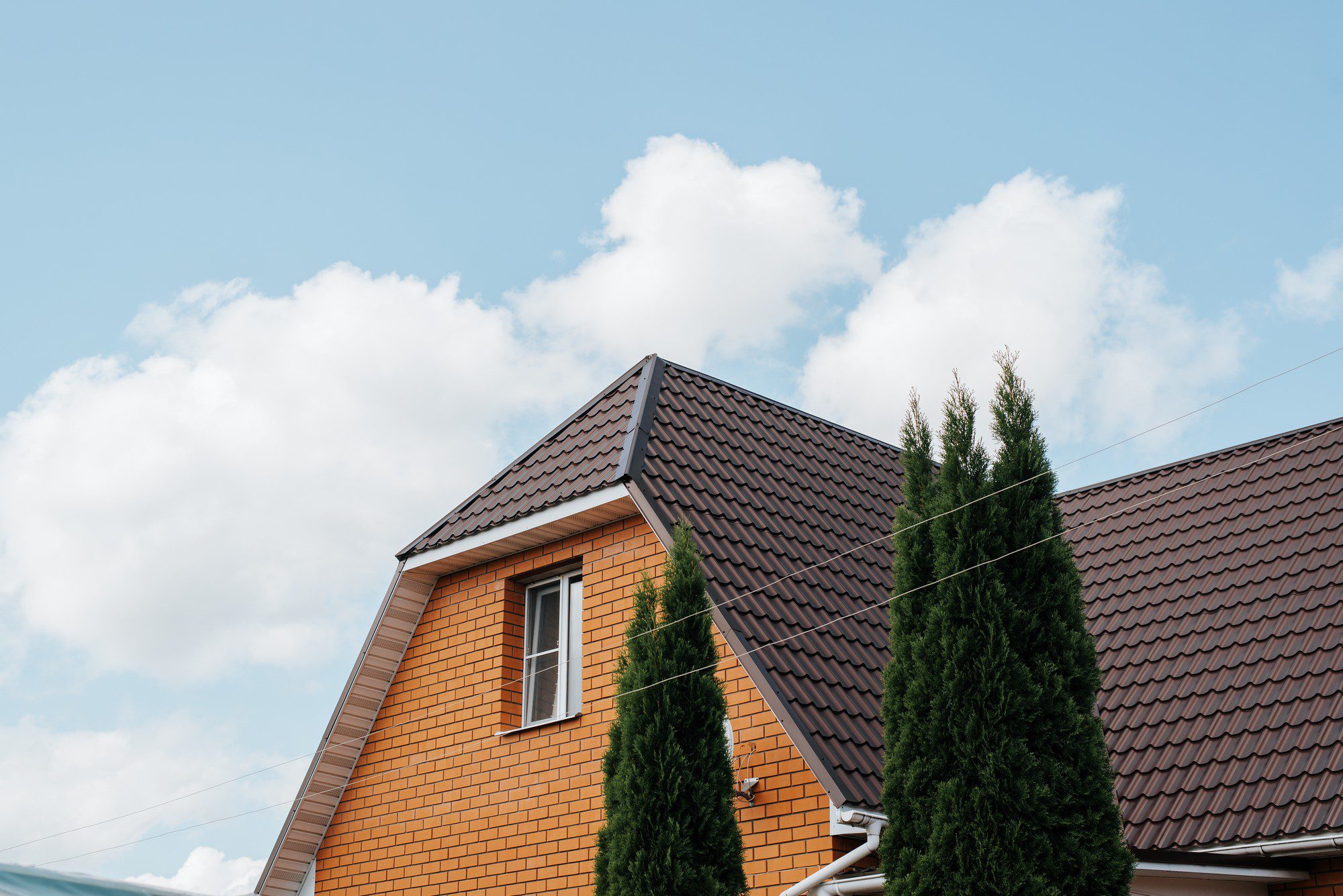 In this image, you see the upper part of a house with an orange brick facade and a dark, tiled roof. To the right of the house, there's a tall conifer tree, possibly a cypress or thuja, based on its shape. There are also power lines visible in front of the house. The background shows a bright blue sky with scattered white clouds. The architecture suggests a residential setting, and the overall weather conditions indicate a clear and potentially pleasant day.