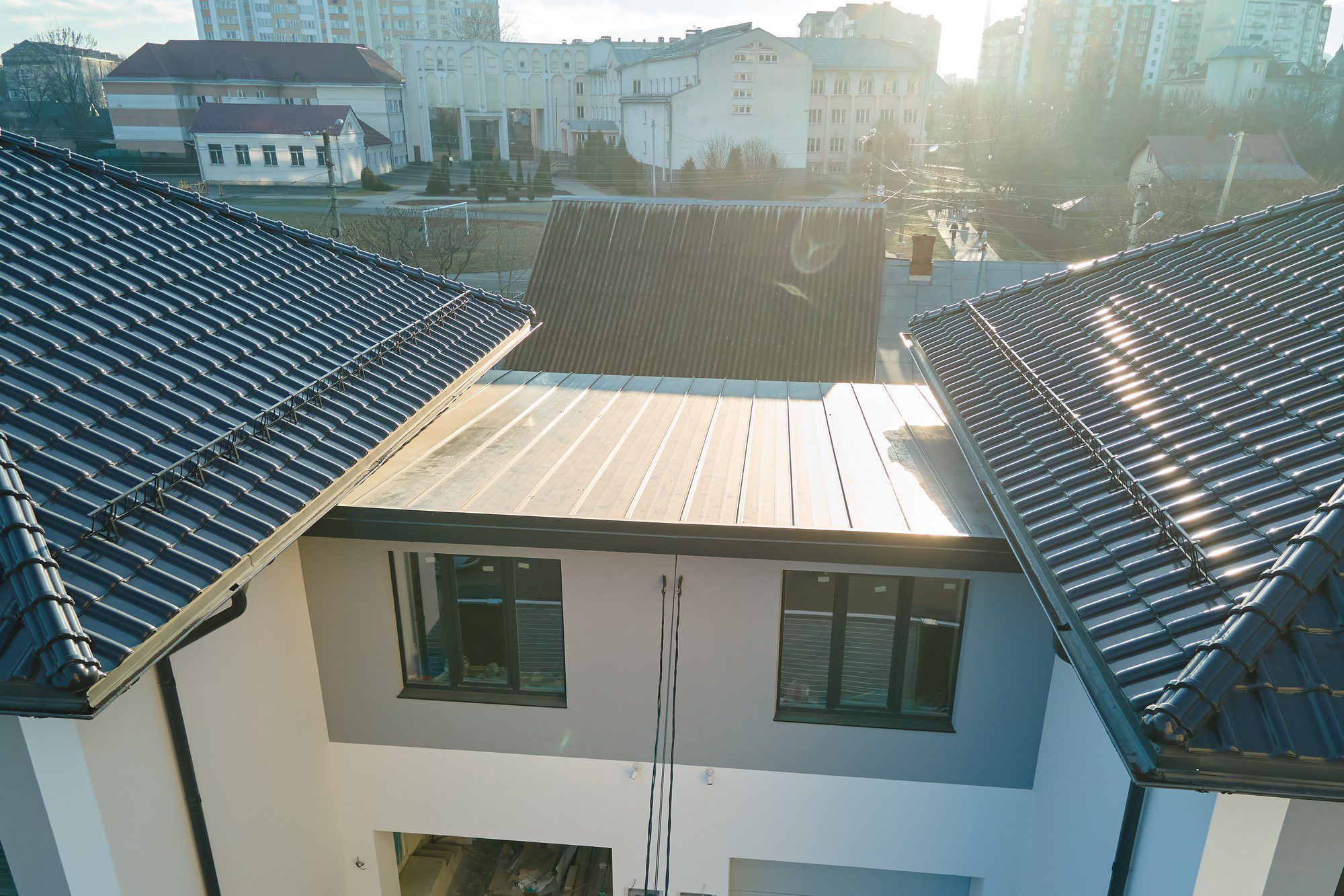 This image shows a view of a modern building's exterior from an elevated perspective, emphasising the roofs. The building features a combination of roofing materials: on the left and right, we see dark roof tiles, possibly ceramic or metal, while the central section has a metal roof with a standing seam profile. The photo captures the sunlight casting a glare and producing lens flare, which indicates that the photo was taken facing the sun. In the background, there are other buildings, both residential and commercial, and some trees, suggesting this is an urban or suburban environment. There is no visible activity on the roofs or visible people in the photograph.