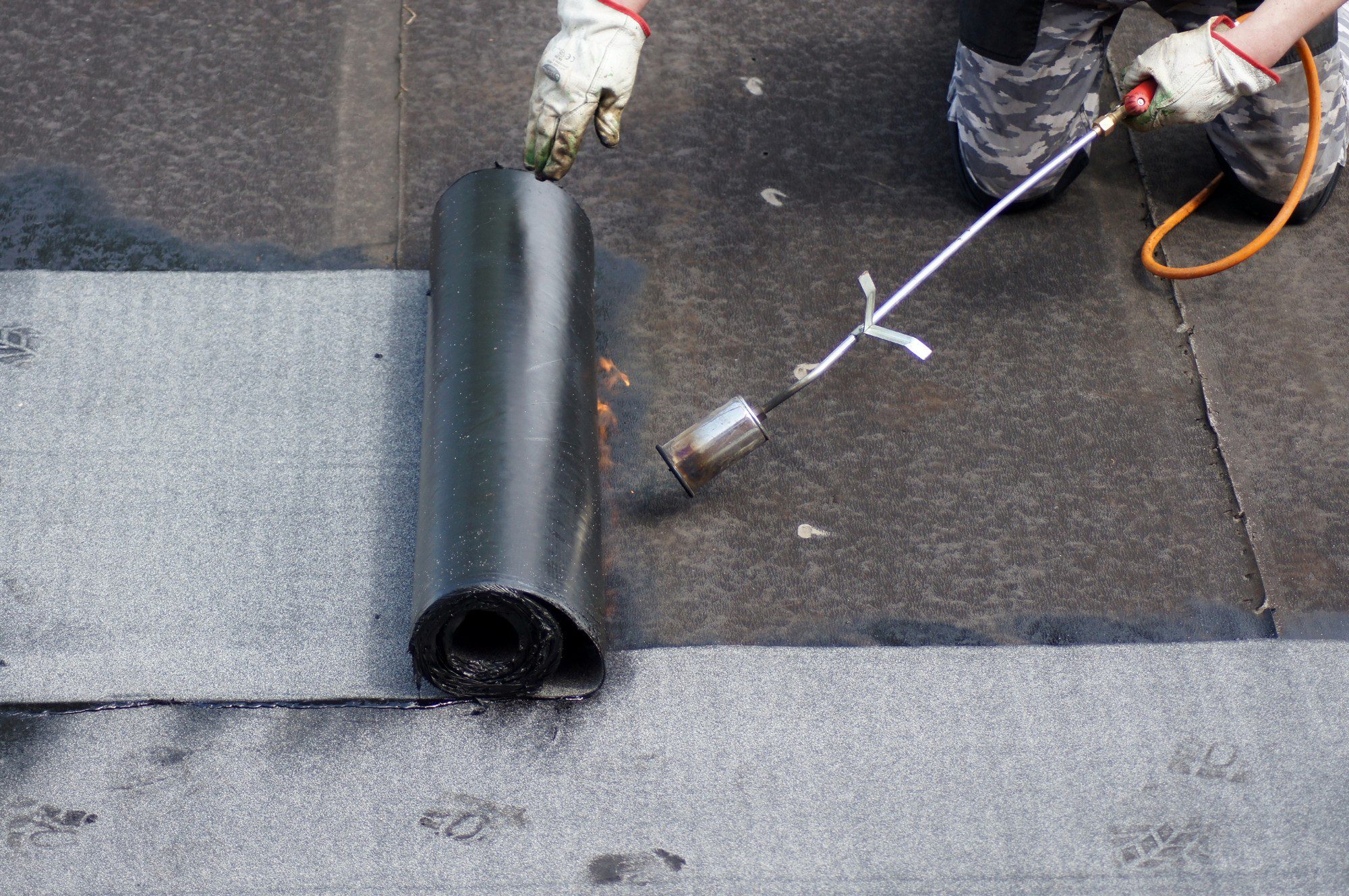 This image shows a roofing process in progress. You can see a rolled-up section of roofing material, likely bitumen-based, which is used for waterproofing roofs. To the right, there is a person wearing gloves holding a torch with an attached hose, which is used to heat the underside of the roofing material. The heating causes the material to melt and bond securely to the roof surface, ensuring a watertight seal. The person appears to be in the process of installing the roofing material on a flat roof. The act of smoothing out and flame-heating the roofing material is part of the application technique for this type of roofing system.