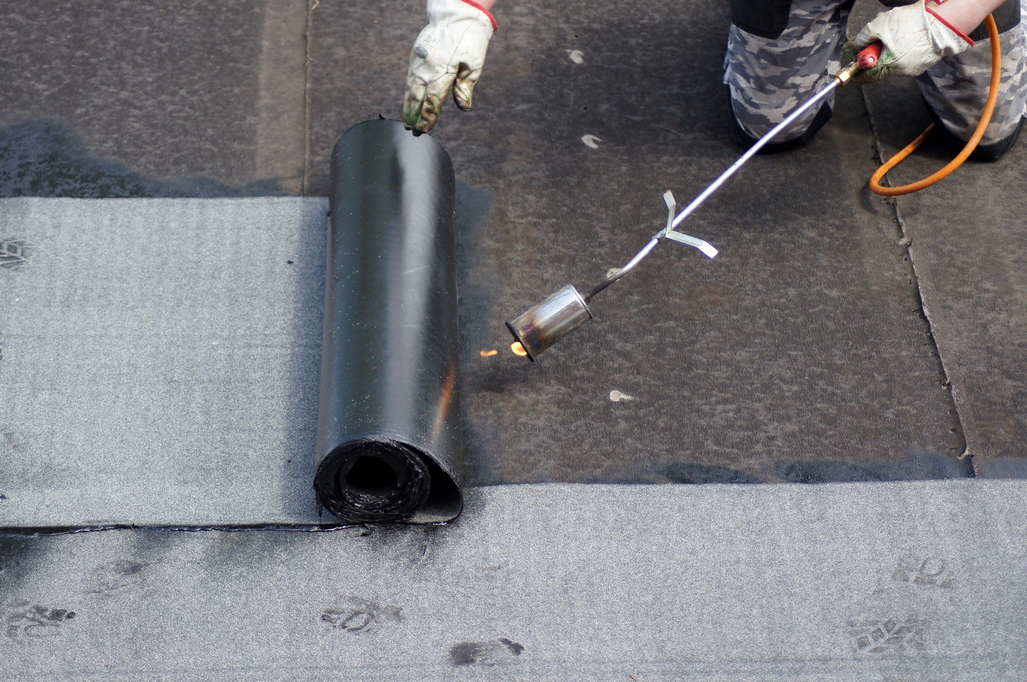 The image shows a roll of roofing material (likely bitumen-based) on a flat roof. A person with a gloved hand is holding the roll, while the other hand holds a propane torch that is heating the underside of the material. This process is typically done to adhere the roofing material to the substrate by melting its underside, creating a bond as the material cools and solidifies. It's a standard procedure in the installation of certain types of flat roofing systems, where a durable and waterproof seal is needed. The person performing the task is wearing partial protective gear and appears to be in the midst of roofing work.