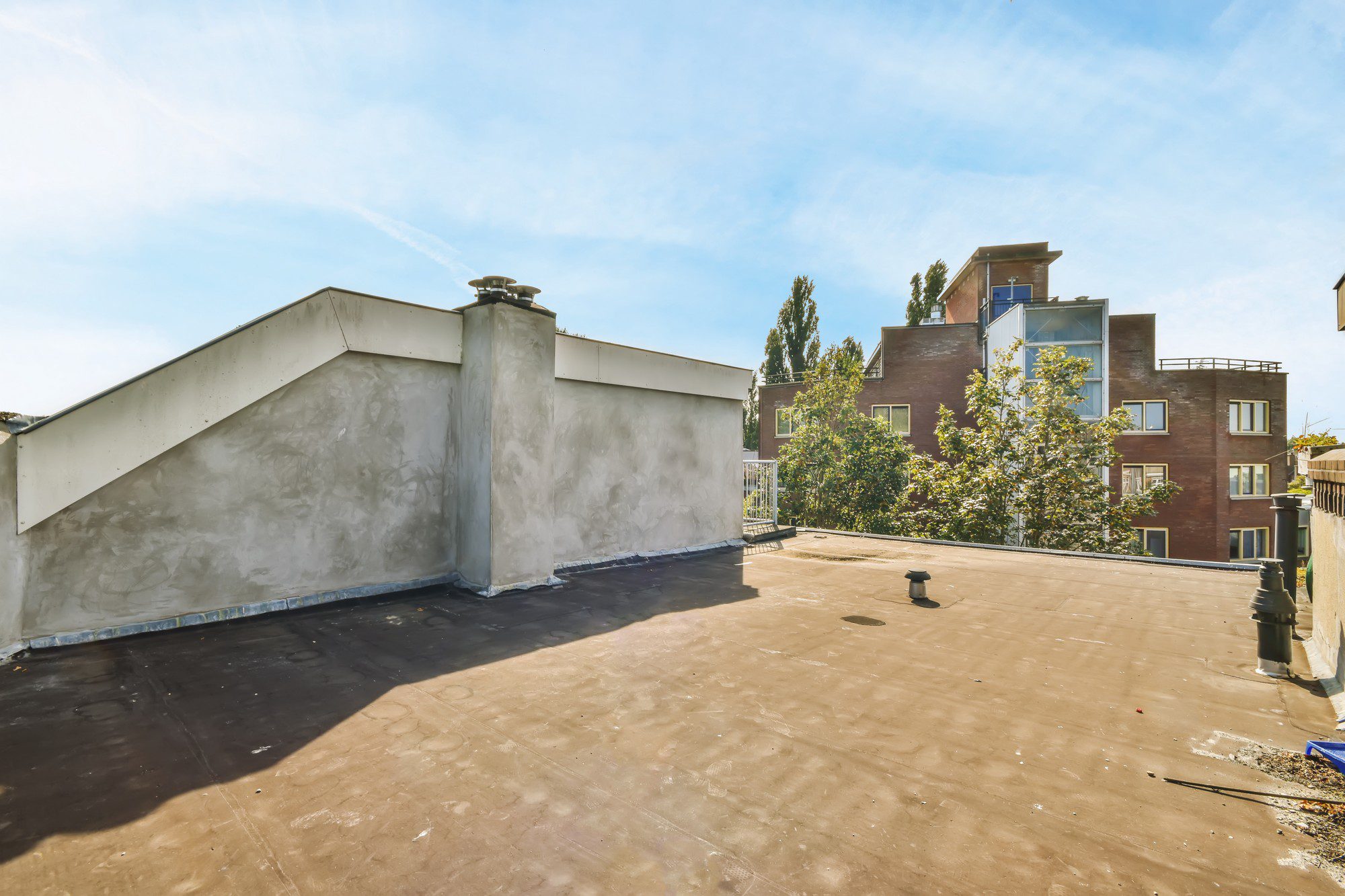 This is an image of a flat rooftop, possibly of an urban flat building. The surface of the roof appears to be some type of waterproof membrane, which is common for flat roofs to prevent leaks. There is a parapet wall surrounding the edge of the roof with a metal capping on top, which provides a safety barrier and is also typical in rooftop designs. A guardrail is visible at one corner, possibly indicating a rooftop access point or patio area. You can also see various protrusions from the roof, such as vent pipes, which are necessary for the building's ventilation system. In the background, there are other urban residential buildings with windows, and a treetop can be seen, adding a bit of greenery to the scene. The sky is clear and it appears to be a sunny day.