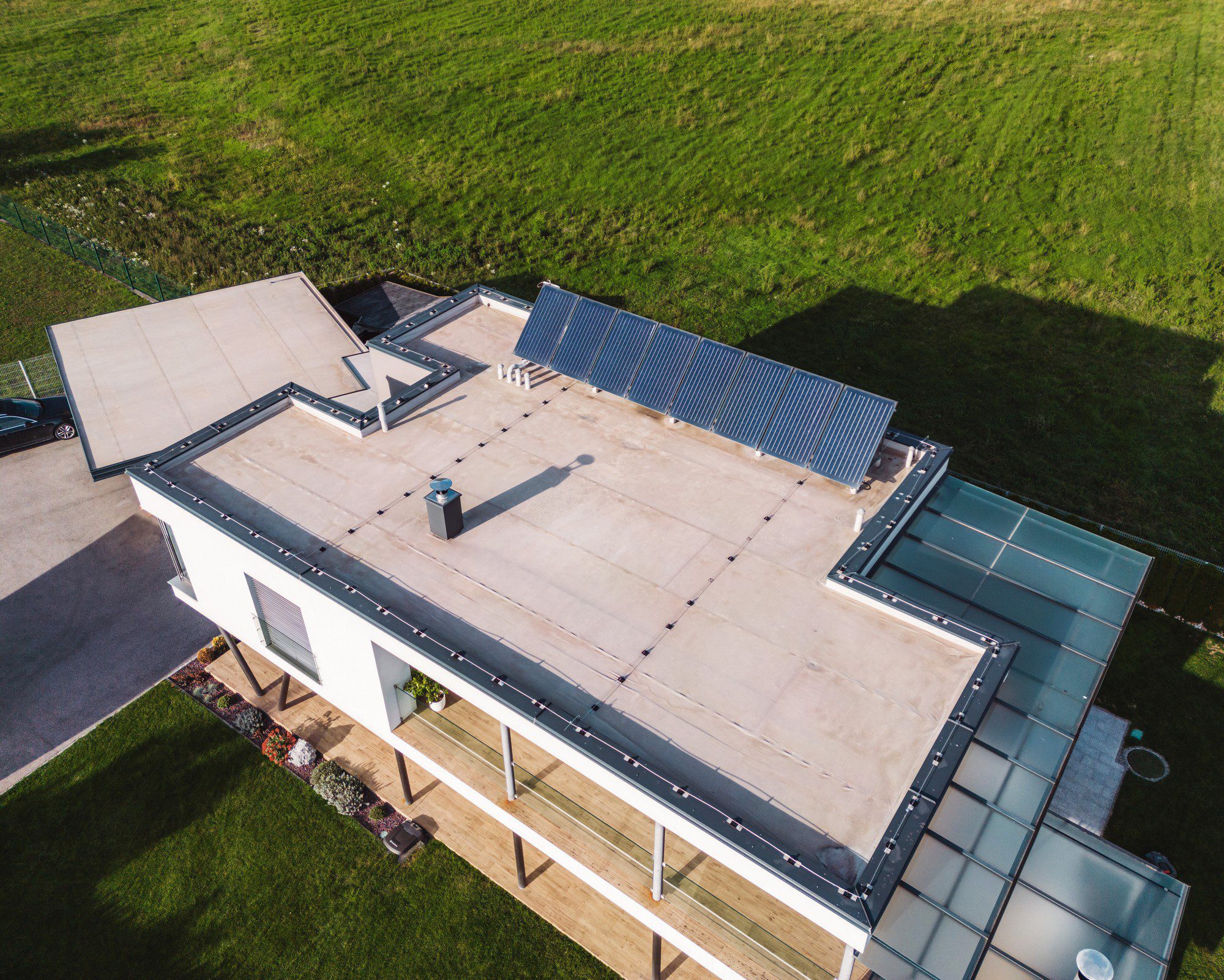 This image shows an aerial view of a modern single-story house. To the left side, on the roof, there's a solar panel installation, which suggests the residents have invested in renewable energy. The rest of the rooftop appears to be flat and is fitted with various roof penetrations, perhaps for ventilation or other services.

Below, you can see a neatly landscaped strip with plants and shrubs alongside the house, which adds aesthetic appeal to the home's exterior. There's also a green field or lawn area extending into the background, giving the property a sense of openness and space. No real people are discernible in this image.