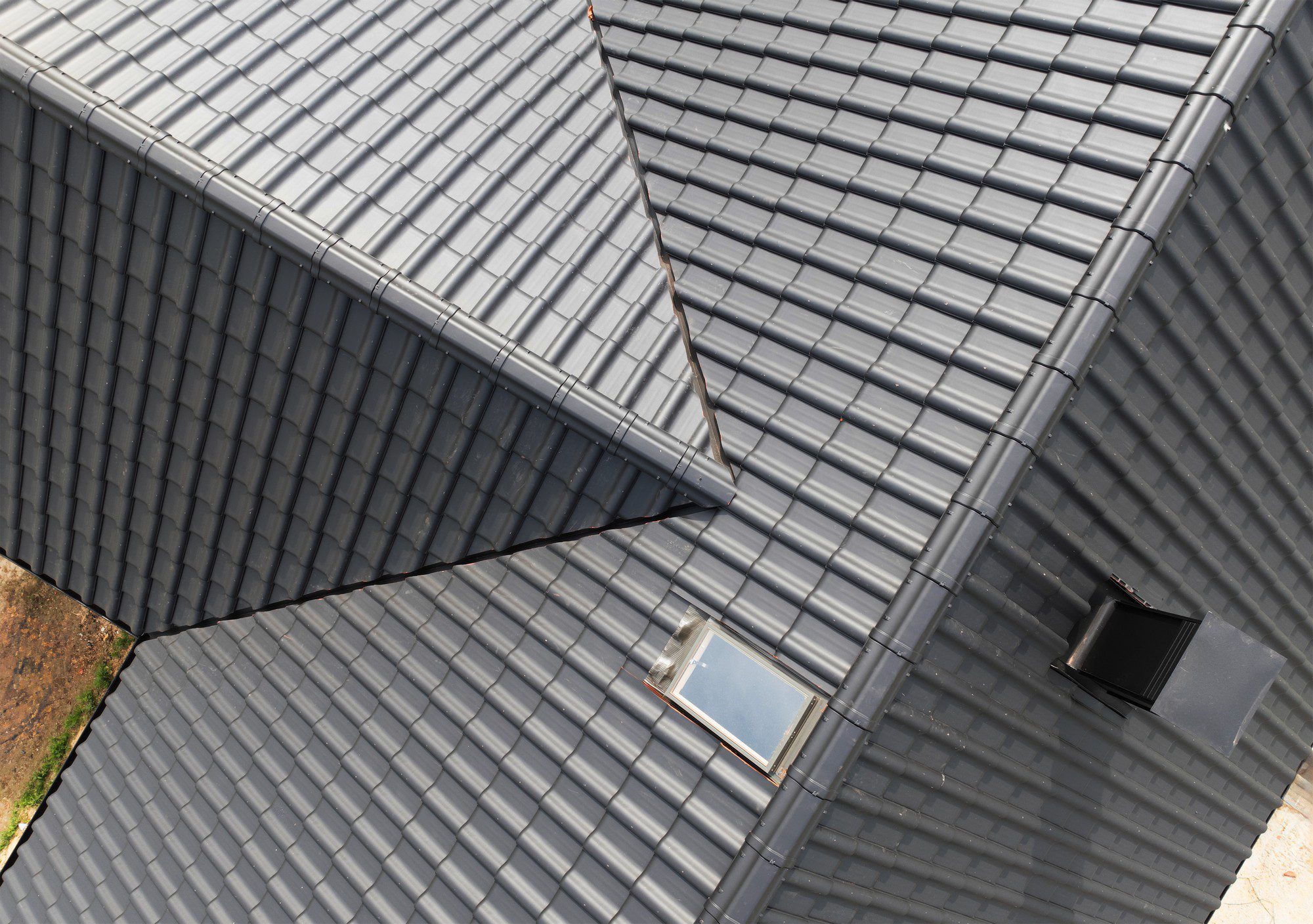 The image shows a close-up aerial view of a roof constructed with gray metal tiles. The composition of the image is geometric, focusing on the intersecting lines and angles formed by the roofing sections. A skylight and a ventilation pipe are also visible, creating a contrast with the uniform pattern of the roof tiles. The skylight allows natural light to enter the space below, while the vent pipe suggests an underlying system for air circulation. There appears to be a slight discoloration or debris on some parts of the roof, which could indicate outdoor exposure. The angle of the photo suggests it was taken from an adjacent higher point, like from another part of the building or using a drone.