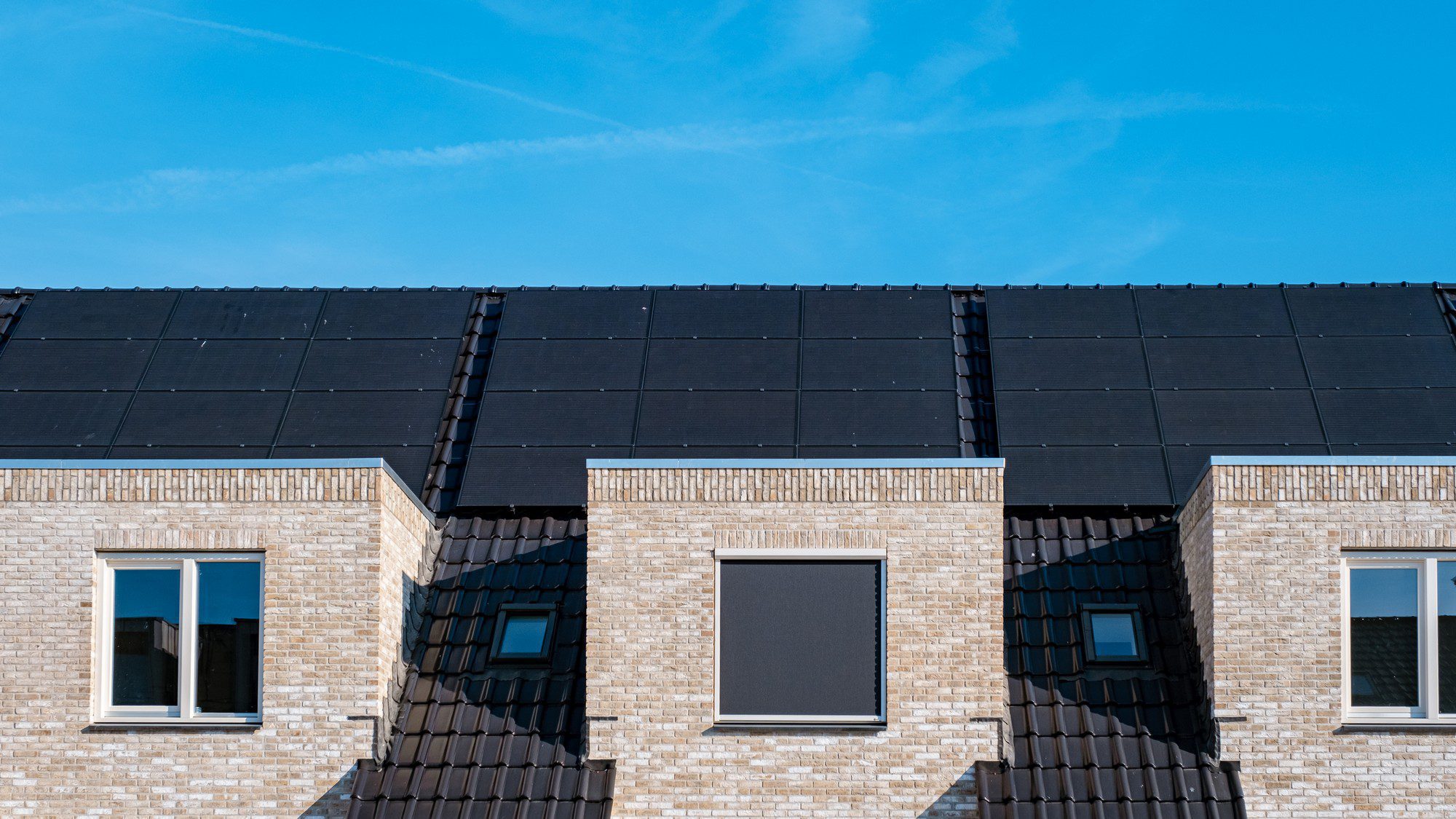 This image shows the upper facade and roof of a brick building with modern solar panels installed on the roof. The sky is blue with minor cloud traces, and the roof has dark, possibly black, roof tiles that complement the solar panels. The panels are arranged neatly across the roof, suggesting an emphasis on energy efficiency and renewable energy use for the building.