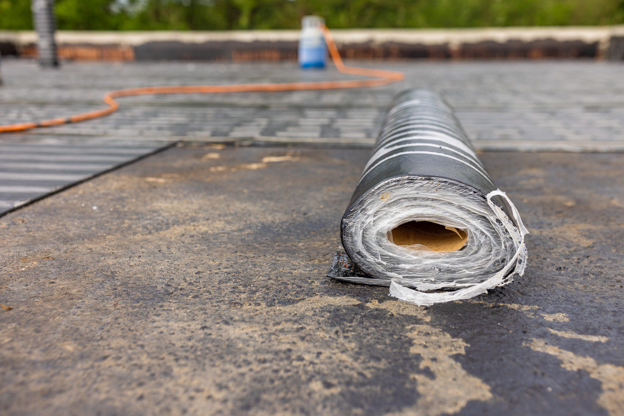 The image shows a large roll of material, likely roofing felt or a similar waterproofing membrane, on a flat rooftop. The roll is partially unwrapped, revealing its layers. In the background, there's another similar roll and an orange extension cord trailing across the roof, leading to a roof parapet. This suggests that some roofing work may be underway or about to start. The top surface of the roof appears to be slightly worn, with some areas where the underlying layer is exposed, indicative of a roof that is being repaired or replaced.