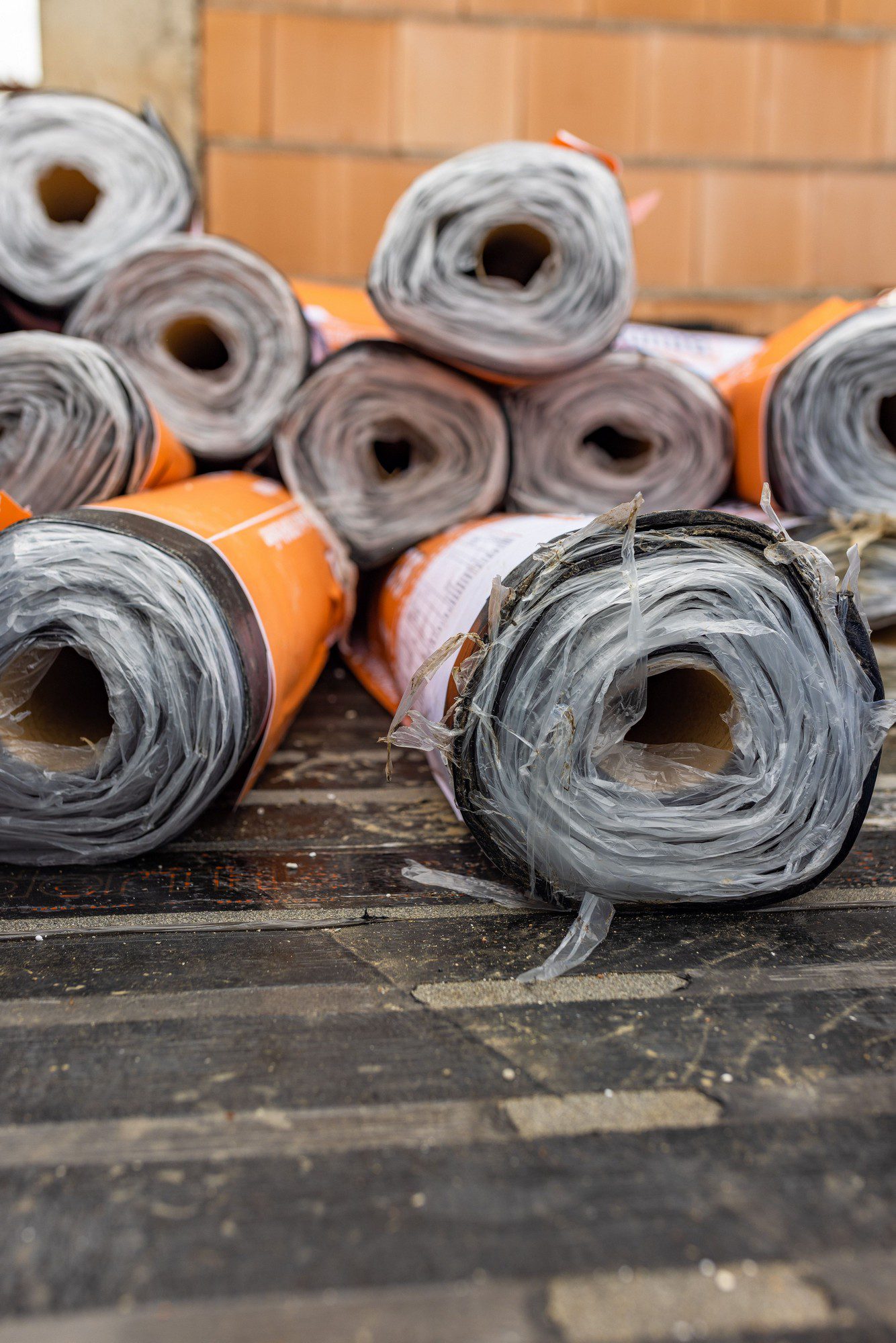 The image shows a number of large rolled-up materials with an orange and white label, likely some form of building or insulation material. These rolls are stacked on a wooden floor or platform, and one of them is in the foreground with its end unfurling. The exact type of material isn't clear from the image, but it could be a waterproof membrane, a roofing underlayment, or a similar construction material used for creating barriers against moisture or for insulation purposes.