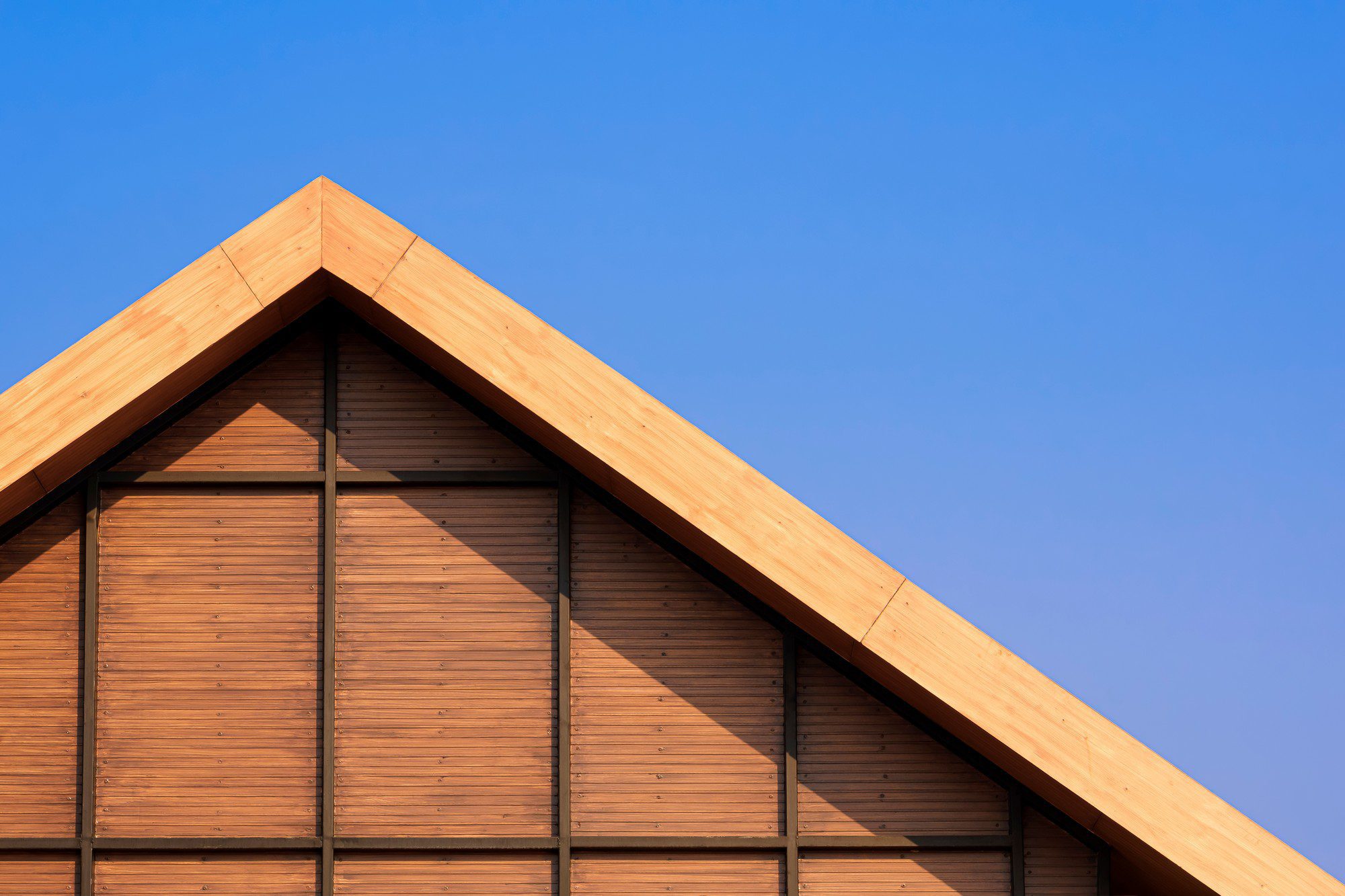 The image shows a close-up view of a building's apex, specifically the gable end of a structure with a pitched roof. The roof and the side of the structure are clad in wood, and there's a clear contrast between the warm tones of the wood and the blue sky in the background. The design is modern and minimalist, with clean lines and no visible roof tiles or other details. The shadow cast on the side of the building forms a triangular pattern, echoing the shape of the roof.